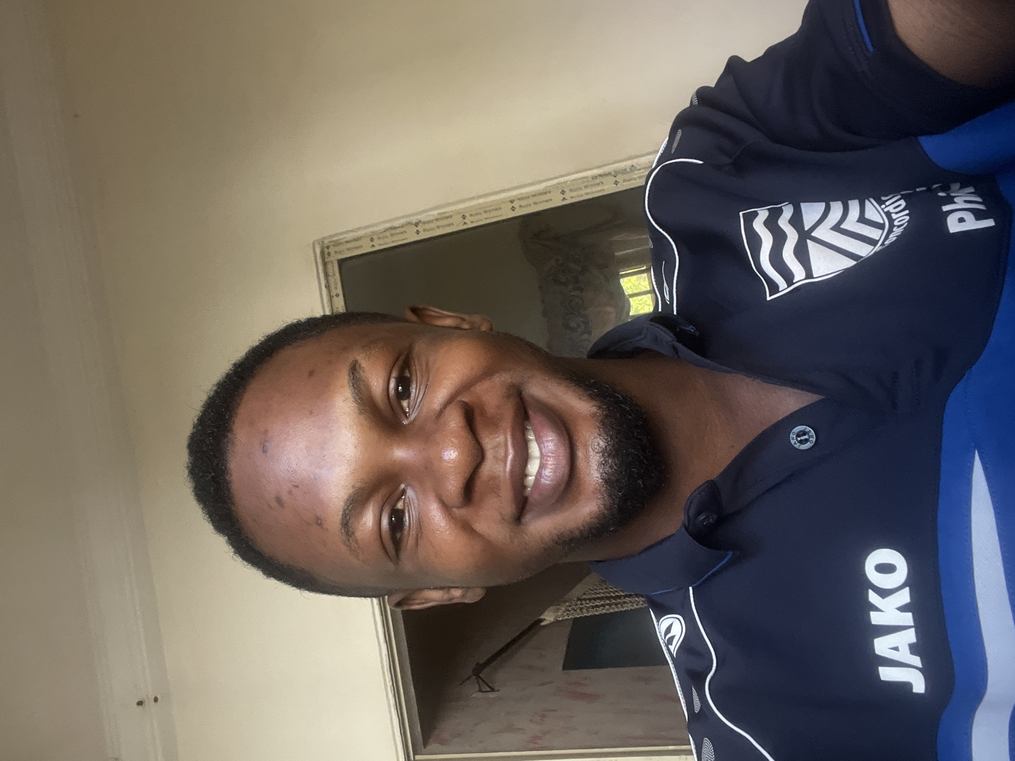A medium shot portrait of a young Black man is captured, taken from a slightly elevated perspective. He is smiling broadly at the camera, his teeth visible. He has short, dark hair and a neatly trimmed beard and mustache. He is wearing a dark blue collared shirt with white and blue trim. The shirt has two logos visible: one on the left chest with the word "JAKO" in white block letters, and another on the right chest featuring a crest with stylized waves and chevron shapes, partially obscured by the collar but with the text "concordi" and "Phi" visible below it.
The man is positioned indoors, with a wall and a doorway visible in the background. The doorway appears to lead to another room and has a light-colored frame. Part of a window with curtains can be seen through the doorway. The walls are painted a light, neutral color, and the ceiling is visible at the top of the frame. The lighting suggests an indoor setting during the daytime, with natural light entering from the right. The overall impression is that of a casual selfie taken within a residential or personal space. There is no discernible evidence to suggest this location is specifically in Jalingo, Nigeria.