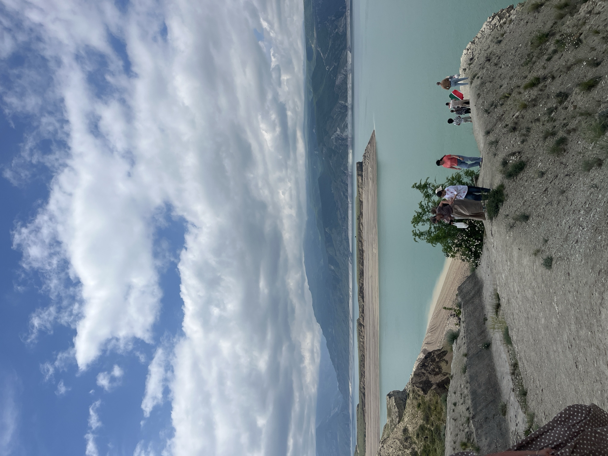 A scenic outdoor shot, rotated counterclockwise, features a cliff edge overlooking a reservoir with several individuals. The sky occupies a significant portion of the left side, displaying a blend of blue and cloud cover.
On the right, a rocky cliff edge serves as a viewpoint, with a group of people standing near the edge, looking out over the landscape. Visible on the cliff, nearest the water's edge, is a tree. Additional individuals are standing further away along the cliff.
The reservoir fills the lower part of the image, exhibiting a light turquoise color, and is flanked by mountain ranges in the distance. A section of the shoreline is visible, appearing as a narrow sandy strip between the water and the mountains.
The scene is likely taken in or around Chirkey, Russia, as indicated by the presence of Chirkey Reservoir.
