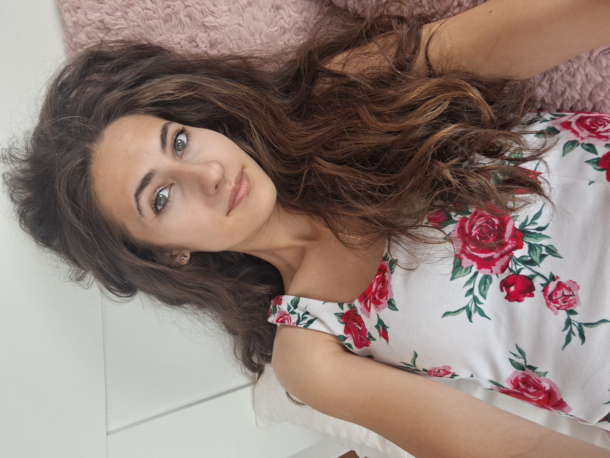 A woman with long, wavy brown hair lies on her back, facing the camera. She is wearing a white tank top with a floral pattern of red and pink roses and green leaves. She has light skin, green eyes, and a light dusting of freckles across her nose and cheeks. Her hair is spread out around her head, resting on a fluffy pink pillow and a white pillow. The background is a plain white wall with horizontal paneling. The lighting is soft and natural, suggesting an indoor setting during the daytime. The woman's gaze is directed slightly upwards and to the left of the camera. Her expression is neutral, with a slight hint of a smile.