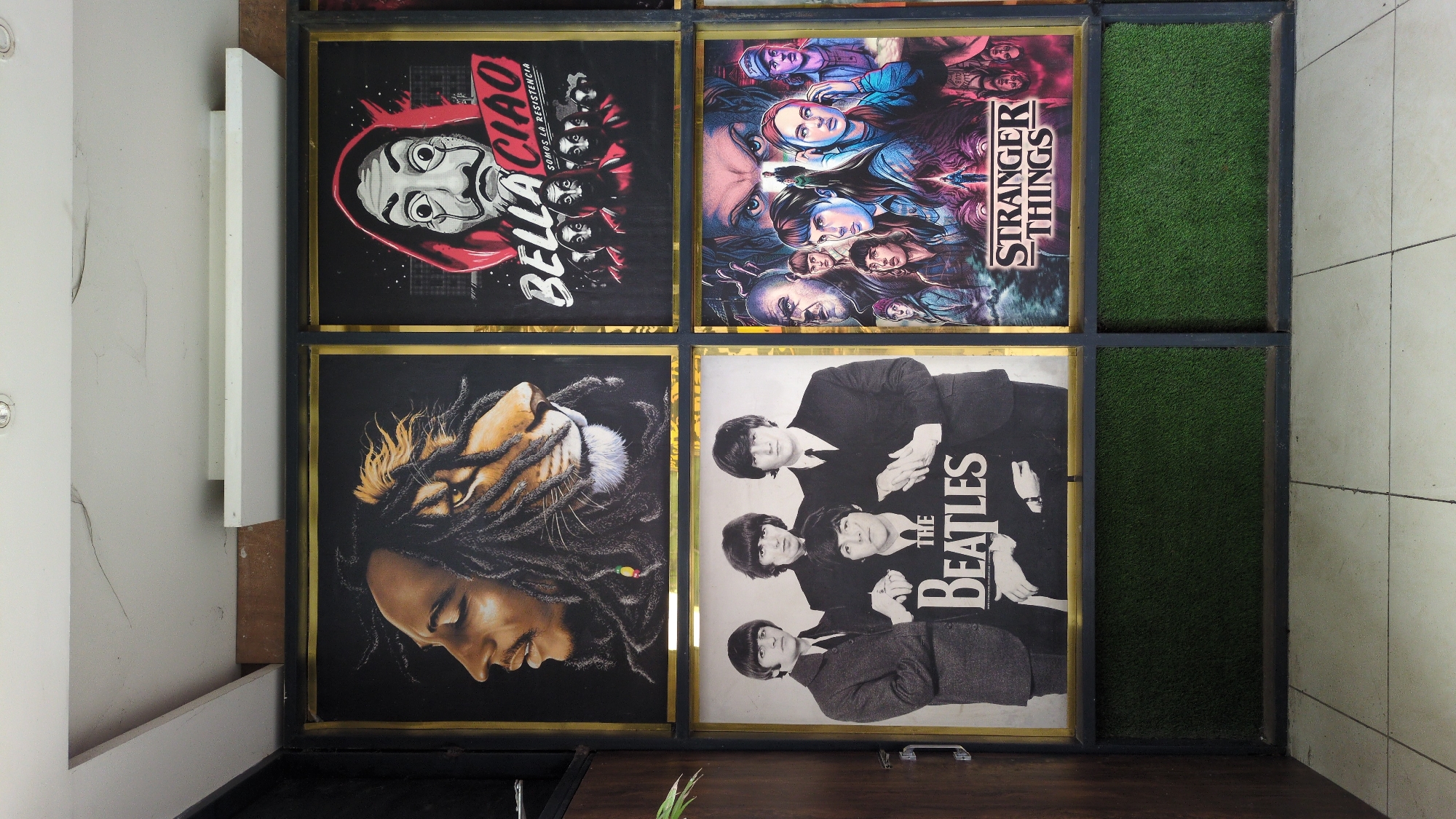 The image shows a collage of posters in frames on a wall. The posters include: "Bella Ciao," "Stranger Things," a portrait of Bob Marley with a lion mane of dreadlocks, and a black and white photo of The Beatles. Two of the six frames contain green artificial turf. The location is Pathankot, India.