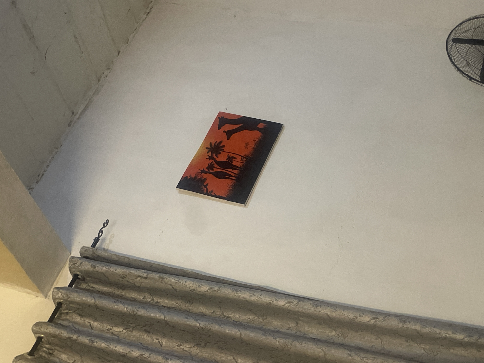 A framed painting of giraffes silhouetted against an orange and red sunset hangs on a white wall in what appears to be a domestic setting. To the left of the painting, a grey patterned curtain is drawn, obscuring a window. The ceiling above is textured and painted a light color. The bottom right of the image shows the top of a black electric fan. The overall impression is of an indoor scene. There is no information in the image to suggest it is located in Jos, Nigeria.