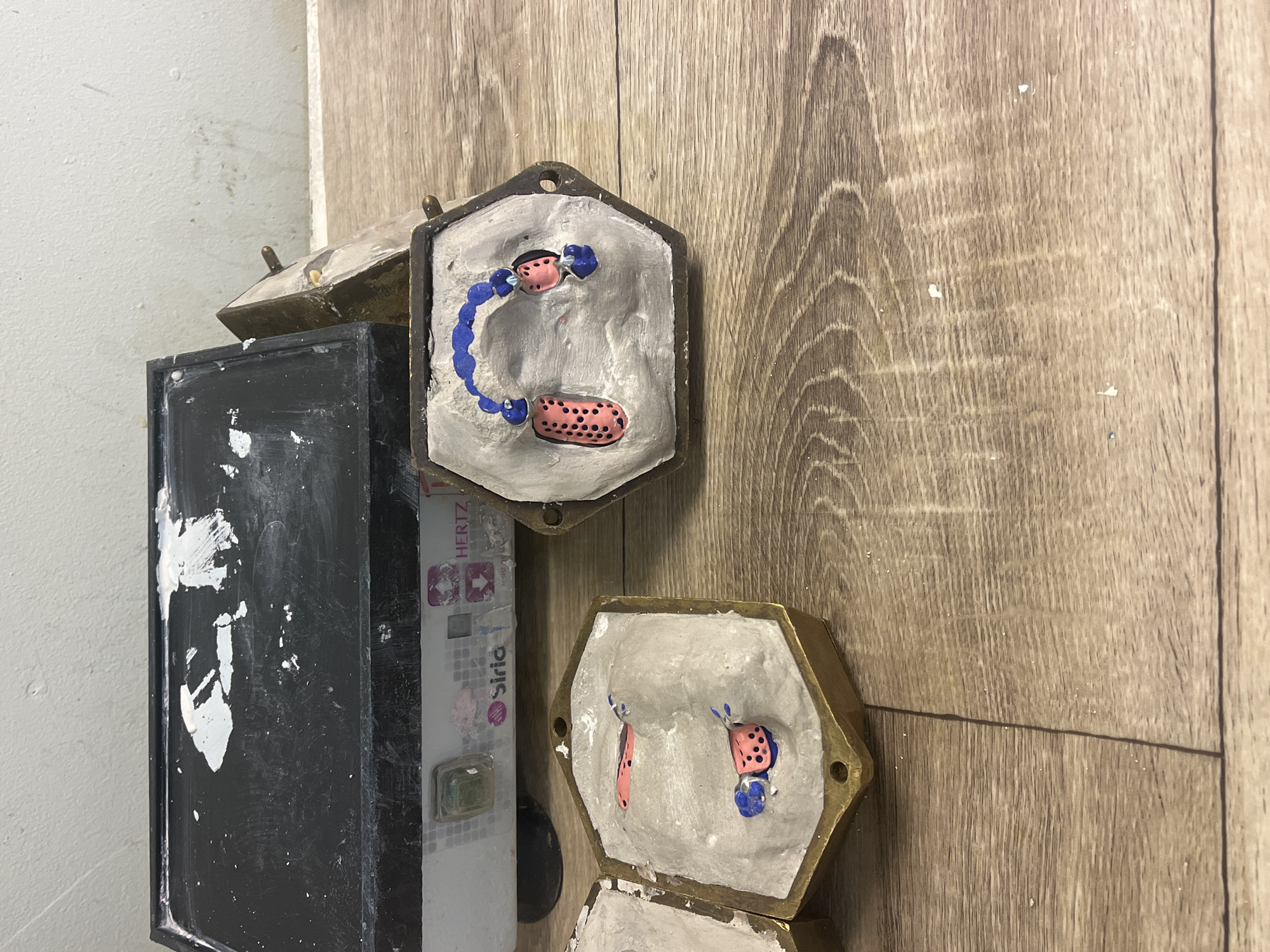 This media file captures an indoor scene, most likely a dental laboratory or workshop. The primary subjects are several hexagonal, metallic molds, some of which are open to reveal their contents.
Inside the open molds, there is a light-colored, plaster-like material. Embedded within this material are elements resembling dental prosthetics. The most prominent mold features a pink, gum-colored section with a stippled texture (black dots), accompanied by curved blue components that appear to be clasps or connectors, forming what looks like a partial denture. Another visible mold below it also contains similar pink and blue prosthetic parts. The plaster inside the molds shows an uneven, cast finish.
To the left of the molds, a tall, black rectangular container or box is visible, heavily stained and splattered with white material, possibly plaster or paint, suggesting an active, hands-on environment. Next to this black box, a white rectangular device is partially seen. Visible text on this device includes "HERTZ" at the top and "Sirio" below it, along with some graphic symbols, indicating equipment or brand labeling.
The background consists of a light-colored wall, showing some signs of wear or dirt, and a wooden surface, possibly a workbench or a wall panel, characterized by its distinct grain pattern.
The scene portrays a functional workspace dedicated to the fabrication of custom dental appliances. There are no people visible, and no specific time of day or weather conditions can be discerned. The overall impression is that of a specialized manufacturing process for dental prosthetics.