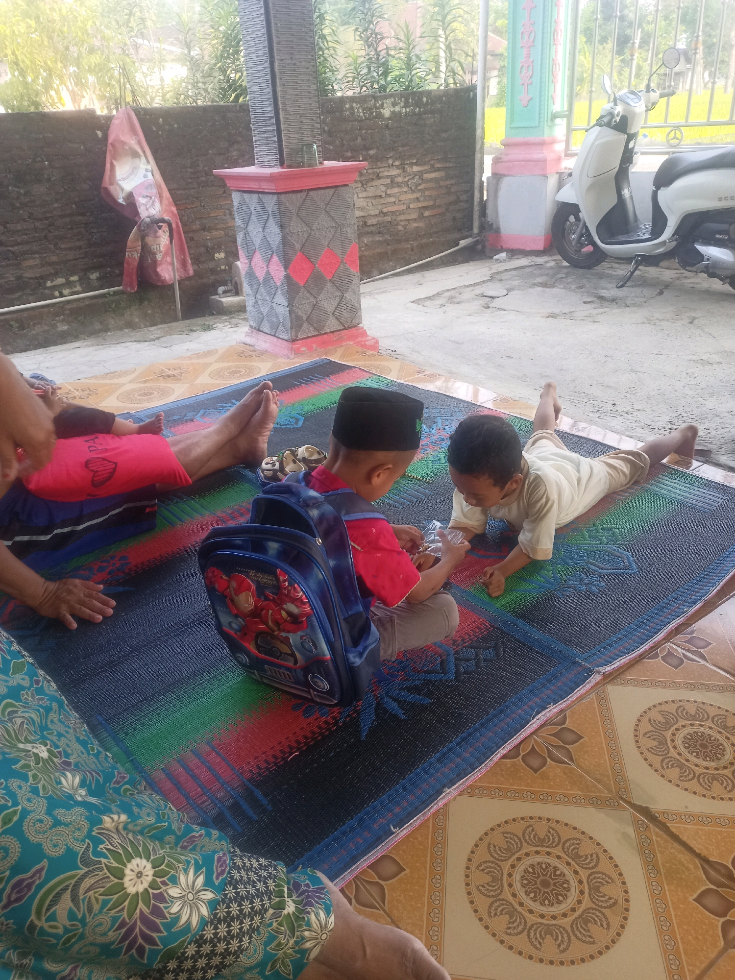 In the outdoor area of Karangtalun, Indonesia, two young boys are situated on a multi-colored woven mat. The boy on the left wears a red shirt and a black peci (a type of cap), and has a blue backpack with an Iron Man design. The boy on the right wears a light-colored shirt and pants, and is lying on his stomach. He is interacting with a small, clear plastic bag. In the background, a white scooter is parked near a decorative pillar and a brick wall. The ground is paved with tiles, and foliage is visible. The lighting suggests it is daytime.
