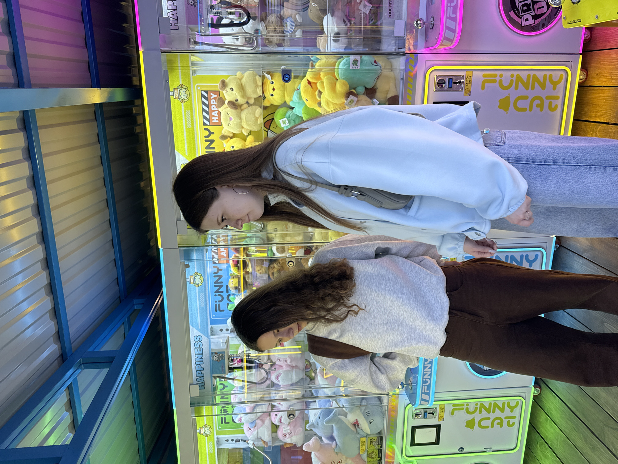 Two young women are depicted in an arcade setting, intently focused on claw machines filled with plush toys. The image is oriented vertically, showing the women standing side-by-side, each observing a different machine.
The woman on the left, with long, straight dark hair, is wearing a light blue jacket or sweatshirt and light-wash jeans. She has a small, light-colored bag slung over her shoulder. The woman on the right has wavy brown hair and is dressed in a light grey sweatshirt and brown pants, with a dark brown bag. Both appear engaged, looking downwards into the machines as if concentrating on a game or selecting a toy.
The claw machines are brightly lit with neon lights in pink, yellow, and green, creating a vibrant, playful atmosphere. The machines contain a variety of stuffed animals, including several yellow Pikachu-like figures, green plushies, and in the lower machine, pink and grey shark toys. The background features corrugated metal panels, possibly part of a ceiling or wall, with blue structural beams.
Visible text on the machines includes "FUNNY CAT" prominently displayed, along with "HAPPY" and "HAPPINESS." Russian text is also present: "КАК ИГРАТЬ" (How to play) and "1 ЖЕТОН - ИГРА" (1 token - game), indicating the cost per play. The scene suggests a casual recreational activity, likely in an entertainment venue in Chaikovsky, Russia.