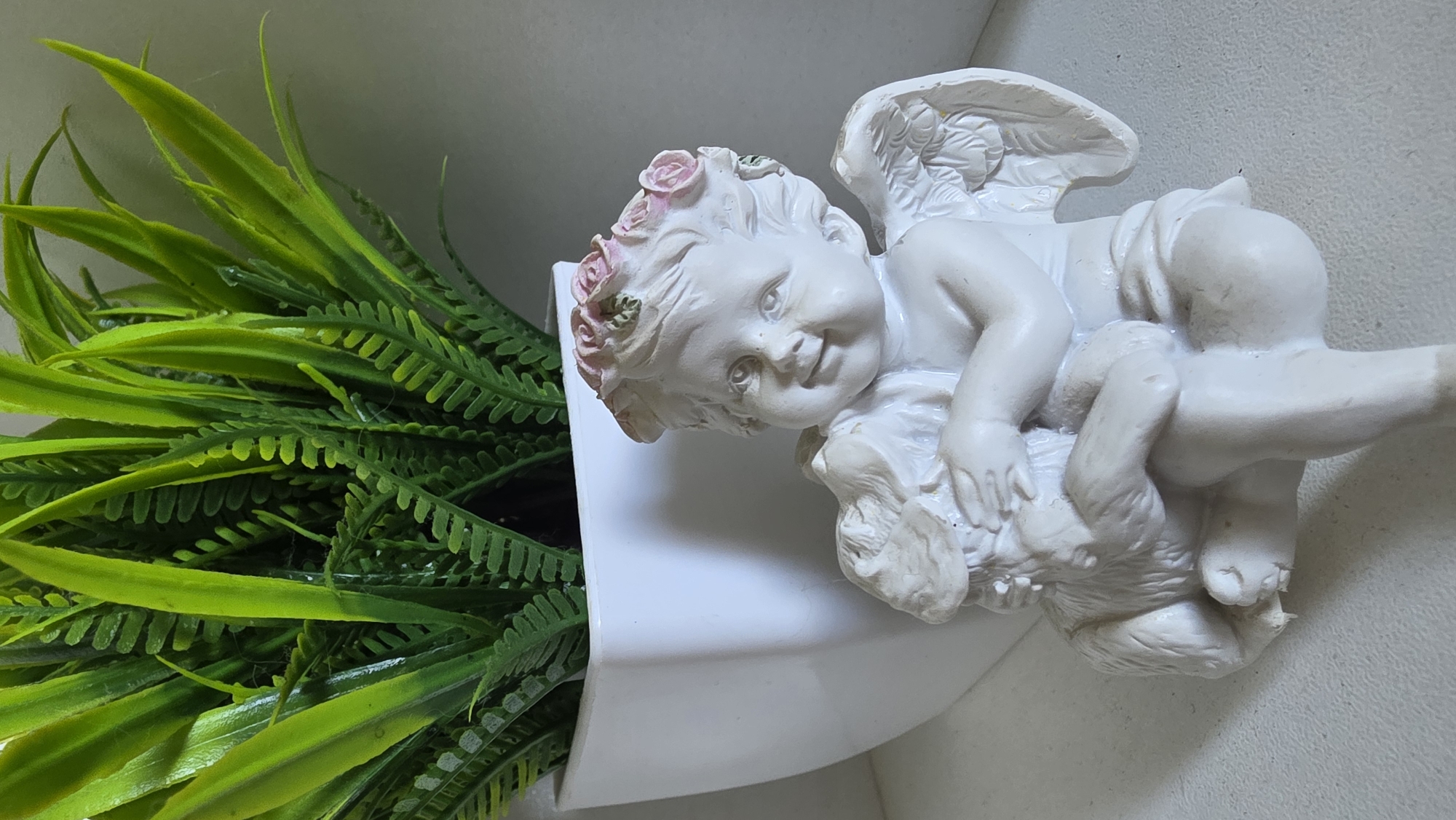 An ornamental white cherub figurine, adorned with a pink floral crown, sits in the foreground, cradling a white animal figurine. Behind it, a white, square-shaped pot holds a lush green artificial plant with long, blade-like leaves. The figurines and pot are positioned on a white, flat surface, possibly a shelf, with a neutral-toned wall as the backdrop. The lighting suggests an indoor setting, likely a domestic environment, as there is no visible indication of the city of Tambov, Russia.