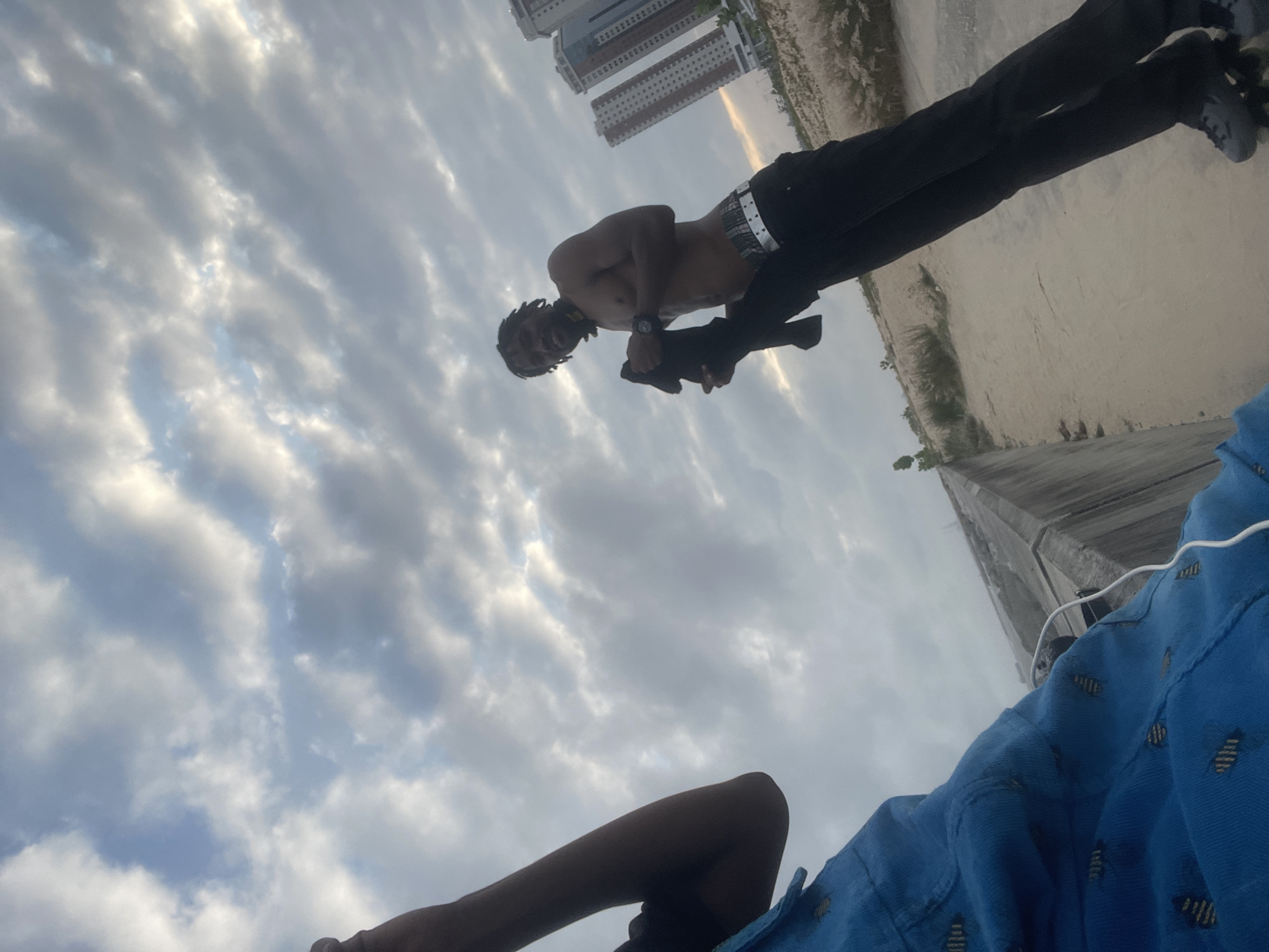 In Inupa Kekere, Nigeria, a young Black male with dreadlocks is pictured shirtless against a cloudy sky. He is holding a black garment in his left hand and wearing black pants with a patterned belt. To his right, three tall, modern buildings are visible in the background. In the foreground, a person in a blue garment, possibly a shirt or jacket with a patterned design, is partially visible. The ground appears to be a dusty or sandy surface, with sparse dry grass. The overall setting suggests an urban outskirts or undeveloped area.