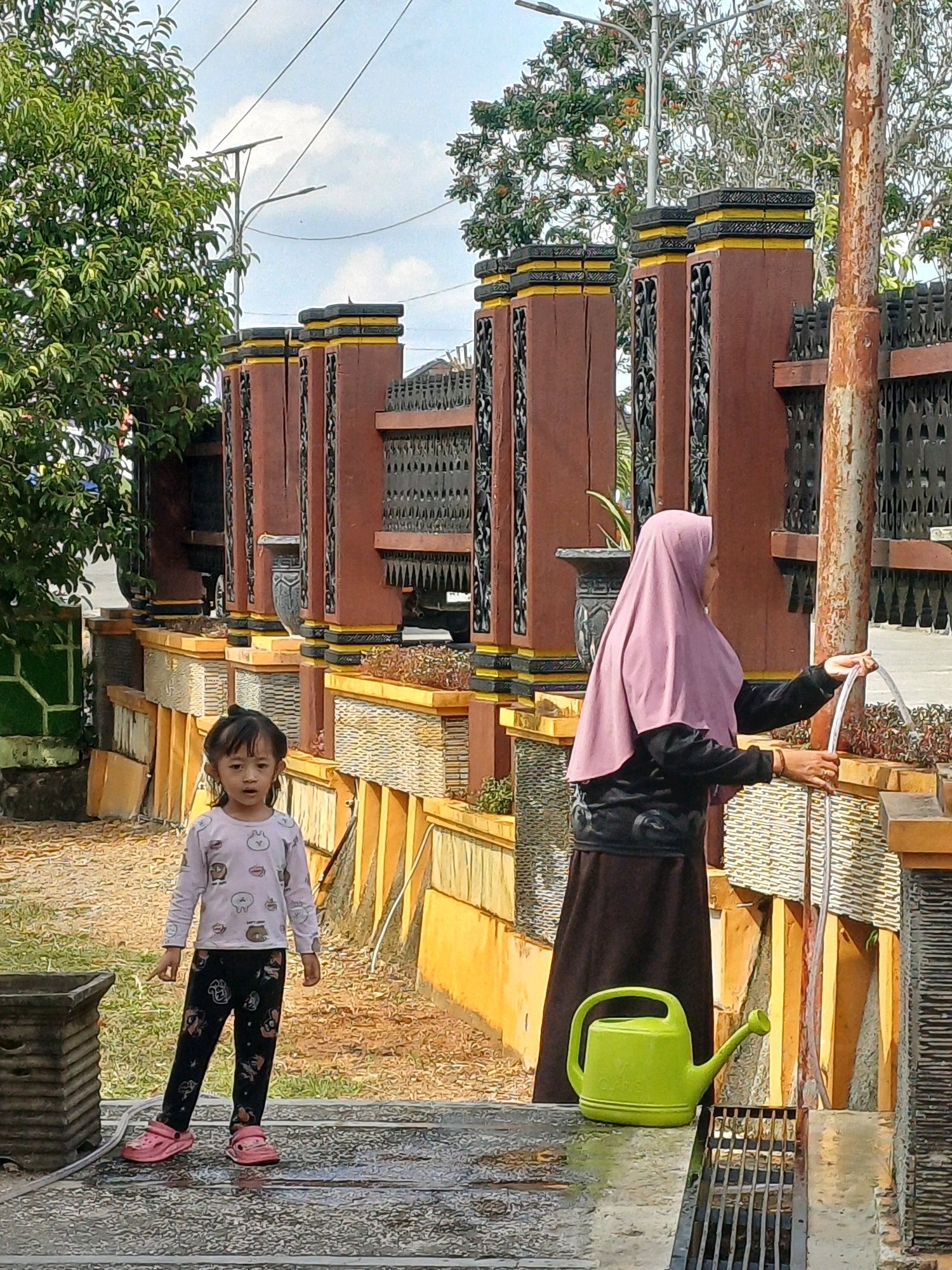 In Pasir Belengkong, Indonesia, a young girl with dark hair stands on a wet, grey surface, wearing a light-colored long-sleeved shirt with cartoon animal prints and black pants with similar patterns. She has pink clogs on her feet. Beside her, a woman in a light purple hijab and a dark outfit is holding a clear hose, directing water into a textured wall. A bright green watering can is placed on a grate to the right of the woman. The background features a decorative fence with brown pillars, yellow accents, and intricate black metalwork. Trees and a street light are visible beyond the fence. The ground is a mix of gravel and water.
