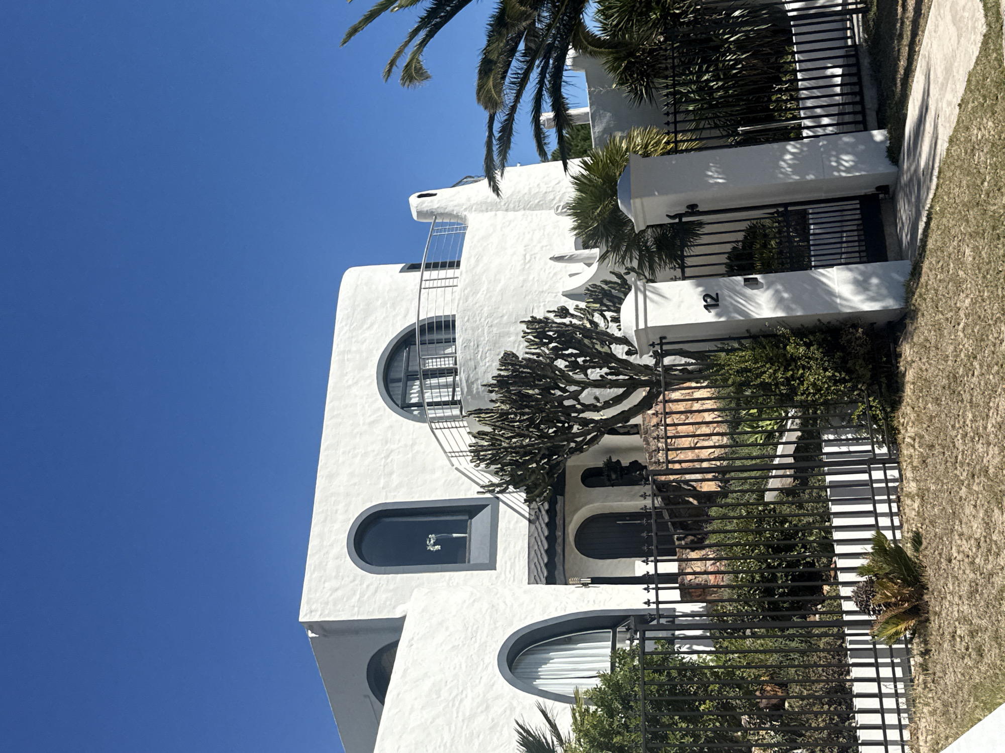 A low-angle, outdoor shot shows a white, two-story building with a distinctive architectural style, potentially located in Cape Town, South Africa. The building's design includes arched windows on both floors and a wrought-iron balcony on the second floor. A substantial, mature cactus or succulent plant is positioned in front of the building, partially obscuring the entrance area.
A dark metal fence runs along the property line, creating a boundary between the building and the surrounding landscape. The fence is interspersed with white pillars. The numeral "12" is visible on one of these white pillars. Grass and vegetation are visible outside the fence. The sky is a clear, vibrant blue. The image is presented in a vertical format, with the building appearing to be tilted to the left. This distortion suggests the image was taken from a low vantage point and possibly at an angle.
