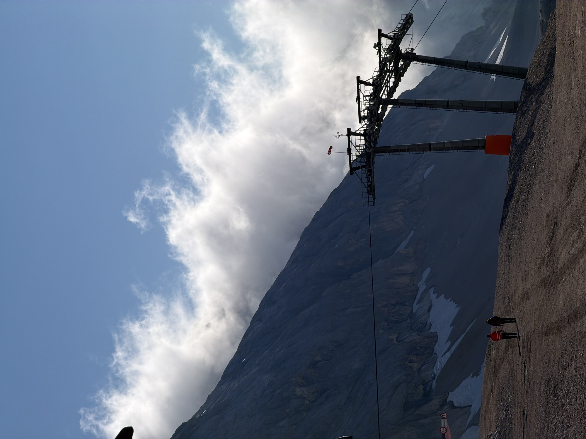 The image depicts a vast mountain landscape under a partially cloudy sky. In the foreground, two individuals walk away from the viewer on a gravelly path. To the right, a ski lift structure with multiple towers and cables is silhouetted against the sky. The mountain slope dominates the background, with patches of snow visible on its dark, rocky surface. The scene appears to be a high-altitude area, possibly a ski resort, given the presence of the ski lift. The sky is a vibrant blue with fluffy white clouds interspersed with wispy formations. There is no specific indication of the city of Garmisch-Partenkirchen, Germany within the image itself, although the mountainous terrain is characteristic of alpine regions.