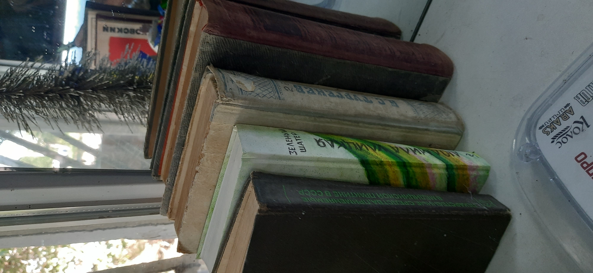 A row of old books is displayed on a windowsill in Dnipro, Ukraine. The books are of varying sizes and colors, with some appearing to be well-worn. The spines of the books are visible, and some have titles and author names written in Russian. The books are arranged in a slightly angled manner, with the one on the left being the largest and darkest in color. Behind the books, a window shows a blurry reflection of trees and a Christmas tree. A small portion of a clear plastic package with text in Russian is visible in the foreground. The overall scene suggests a personal collection of books, possibly in a home or an office setting in Dnipro.