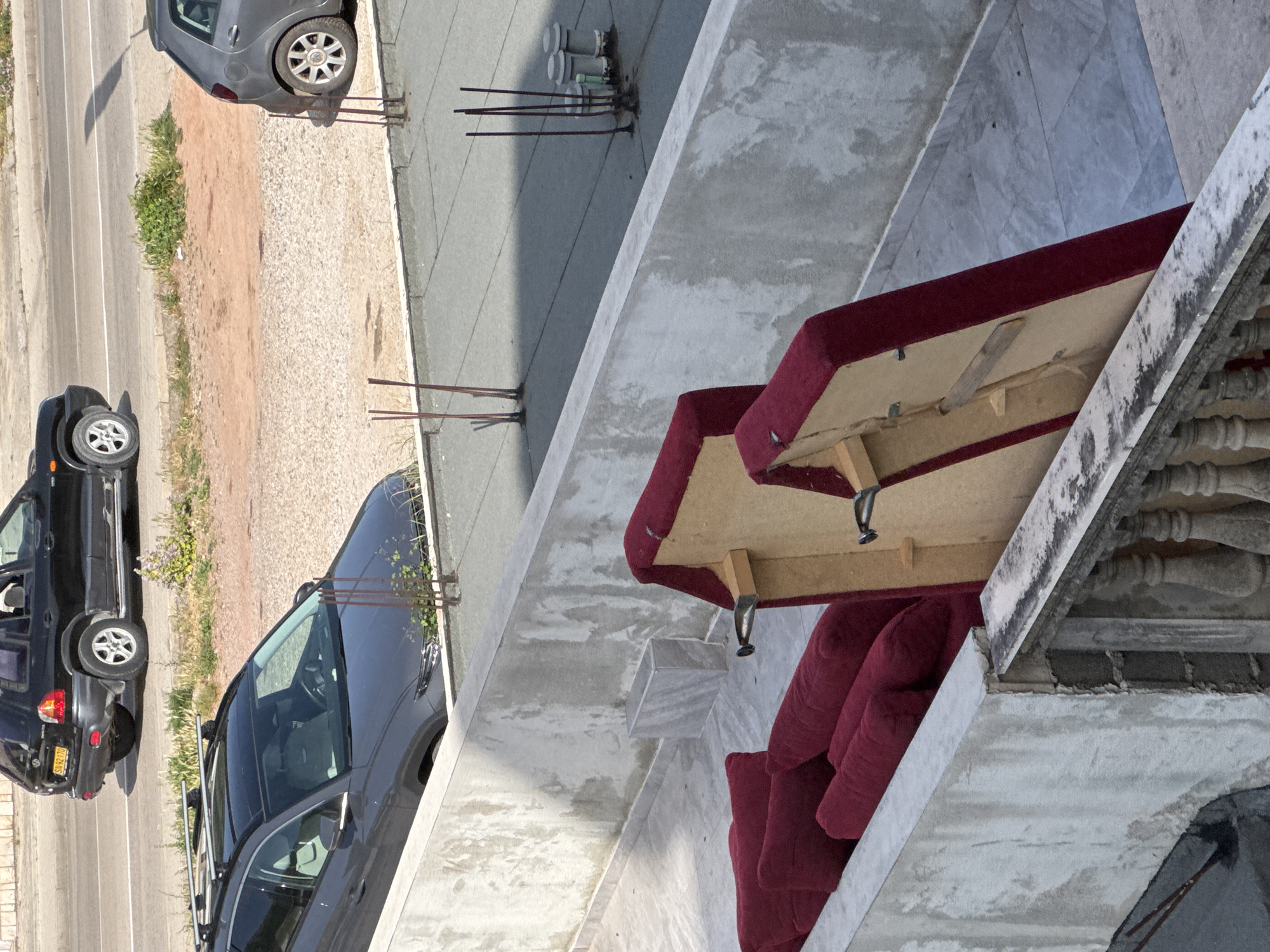 From an elevated perspective, the image captures a view from a balcony overlooking a street in Zankovići, Montenegro. Parked cars are visible on the street, including a black hatchback and a silver SUV with a roof rack. On the balcony, a disassembled red sofa is visible, with the base upside down, revealing its wooden frame and metal attachments. The balcony itself has a weathered concrete structure with a decorative railing. The overall environment suggests an urban or suburban setting.