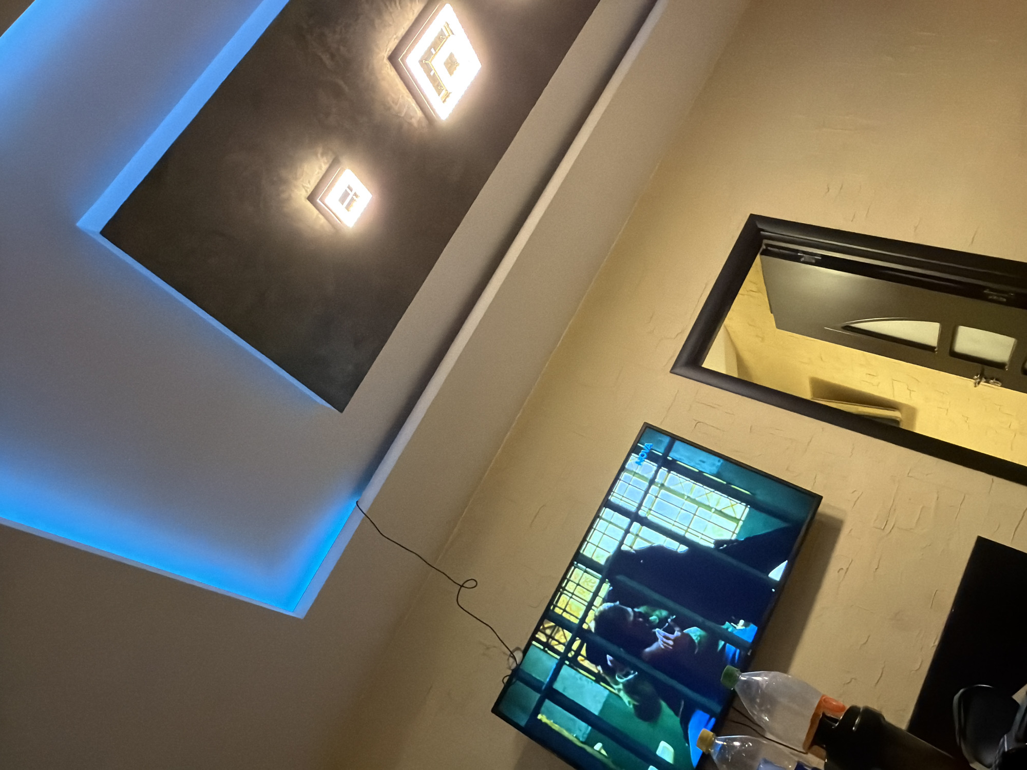 This image depicts an indoor domestic environment, likely a living room or bedroom, located in Plovdiv, Bulgaria. The walls are painted in a textured, light beige or pale yellow hue. The ceiling features a multi-level dropped design: an outer white section with prominent blue LED strip lighting running along its perimeter, providing an upward glow, and an inner, recessed section with a dark, possibly textured grey or brown finish. This darker section contains two illuminated square-shaped ceiling lights, emitting bright white light.
A flat-screen television is mounted on one of the beige walls, angled slightly. The screen displays a scene from a video or broadcast, showing two figures (presumed human) positioned behind vertical bars or a gridded window-like structure. A black electrical cable, coiled in places, extends from behind the television upwards along the wall and into the ceiling. Below the television, a dark, flat surface holds two plastic bottles: one with an orange cap, appearing partially full, and another with a yellow cap, appearing fuller. To the right, a dark-framed doorway stands partially open, revealing a glimpse of another interior room with similarly toned yellow walls. The door itself is dark with distinct paneling and a visible metallic handle/lock mechanism.