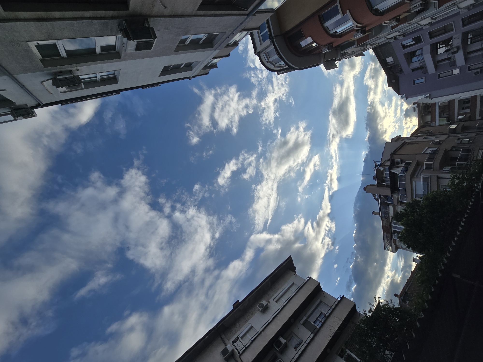The image presents a worm's-eye view looking directly upward at a vibrant blue sky, framed by the surrounding facades of urban buildings.
The central subject is the expansive sky, filled with scattered white clouds of various types and densities. Some clouds appear wispy and elongated, like cirrus or altocumulus, while others are denser and puffier, resembling cumulus clouds. A striking feature is the presence of distinct sunbeams, or crepuscular rays, which pierce through a denser cloud formation in the upper right portion of the sky, indicating the sun's position and bright illumination. The overall sky color is a deep azure.
The sky is encircled by the tops of several multi-story buildings, suggesting a narrow street, alley, or courtyard setting in a city. These buildings are primarily residential or commercial, displaying a mix of architectural styles. Their exteriors feature neutral tones such as light gray, beige, and muted white, with some structures exhibiting accents of reddish-brown and a subdued purple hue. Numerous rectangular windows, some with closed blinds or reflective glass, are visible, along with balconies and railings. Many of the buildings also have external air conditioning units mounted beneath windows or on their walls. Dark green foliage from trees or bushes can be seen peeking into the frame from the bottom and right edges.
The time of day appears to be daytime, likely late afternoon, given the prominent sunbeams and the quality of light. There are no people visible, nor any specific event or activity taking place. The scene is static, focusing on the architectural enclosure of the urban space contrasted with the open, dynamic sky. No visible text is present in the image.