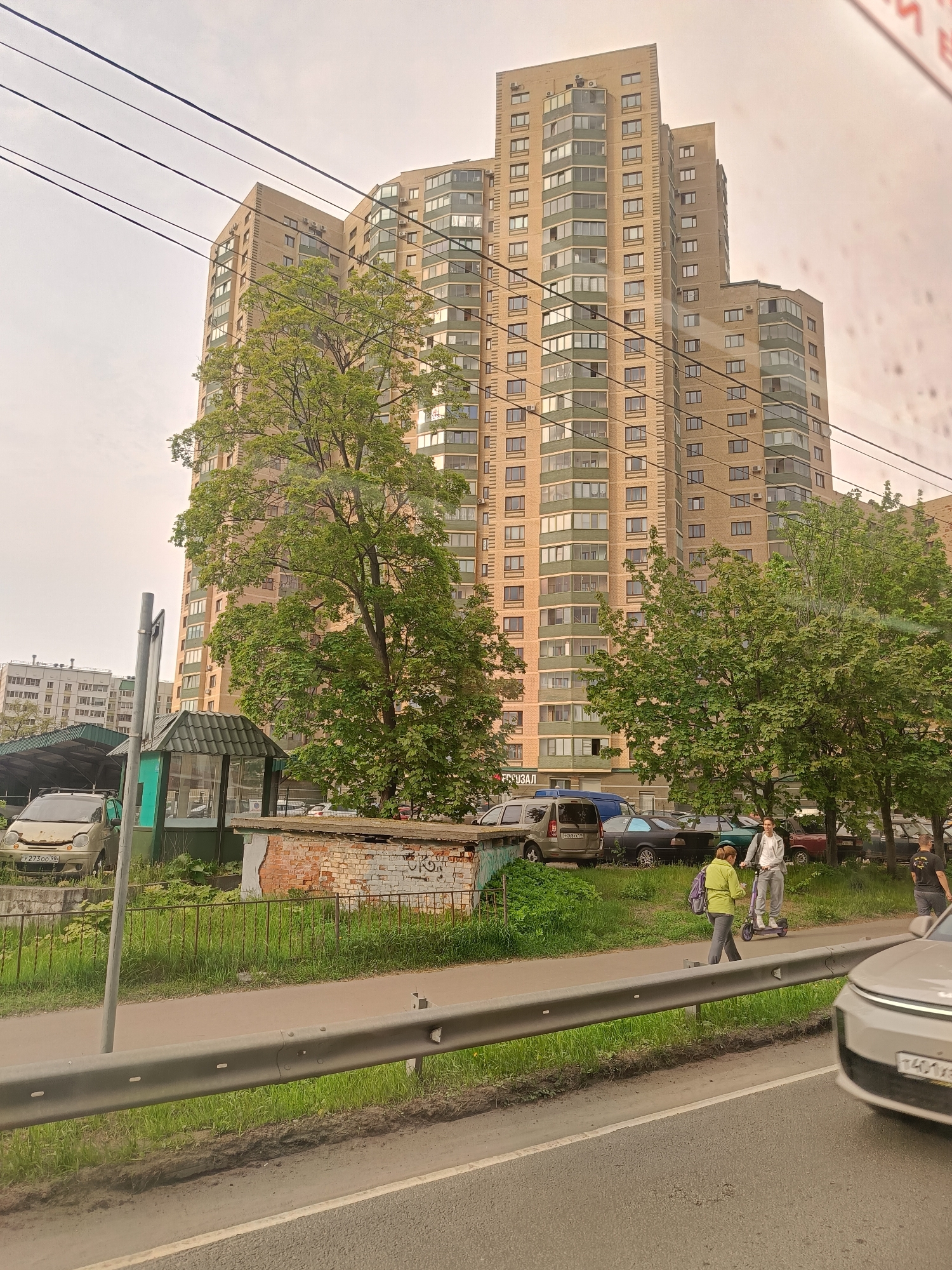 The image depicts a street scene in Saint Petersburg, Russia. A tall, light-brown apartment building serves as the backdrop. In the foreground, a pedestrian walkway is separated from the road by a metal guardrail. A woman in a green jacket walks along the sidewalk, while another person rides an electric scooter. A silver car is partially visible in the bottom right corner of the frame. Behind the sidewalk, a parking area contains several cars of varying colors and models. A small, derelict brick structure with white graffiti is visible next to a green bus stop. A damaged beige car is parked next to the bus stop. Overhead electrical wires crisscross the sky. The weather appears overcast or hazy.