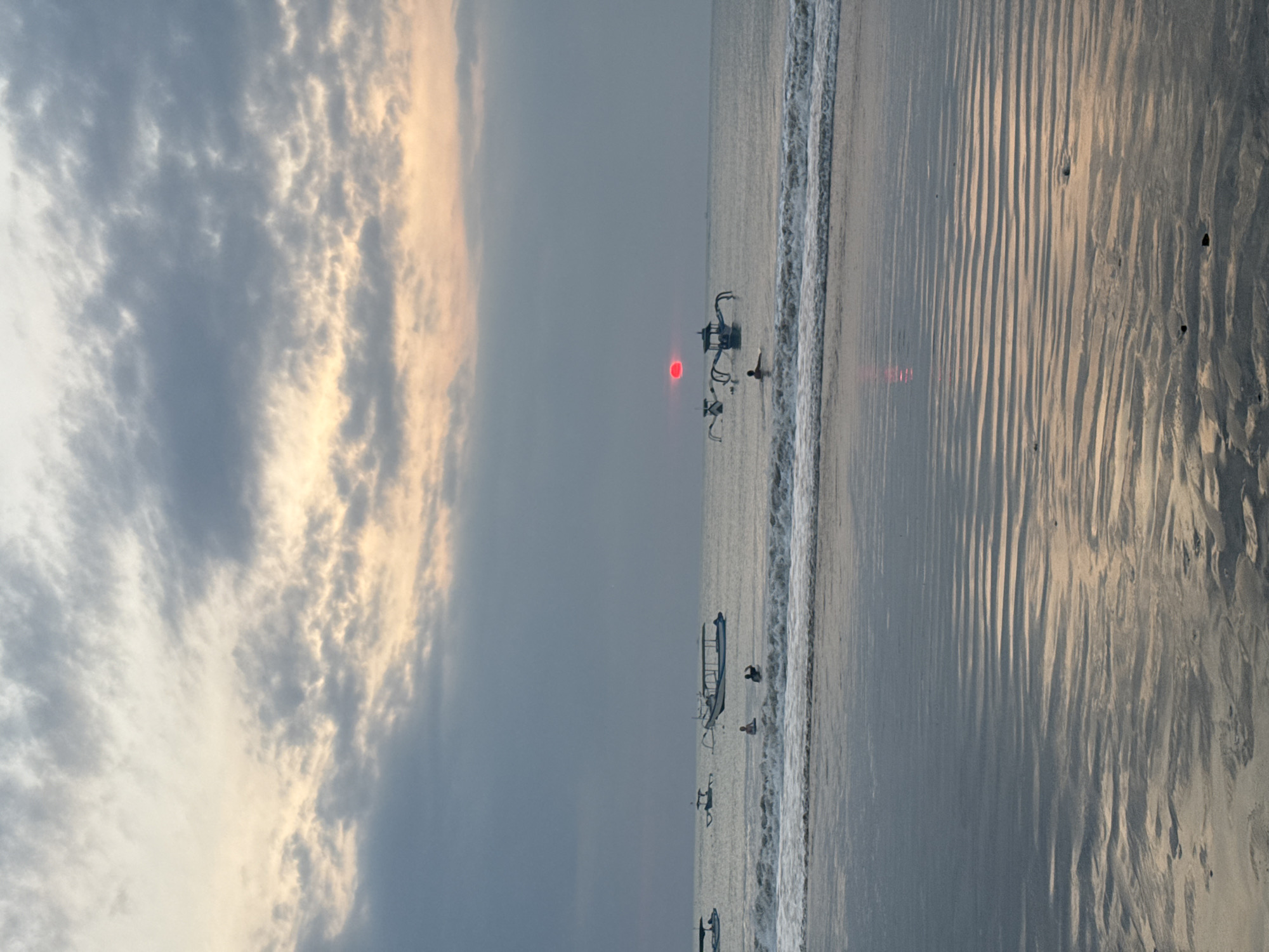 The image depicts a serene beach scene at dusk in Kuta Selatan, Indonesia. The sky is a canvas of muted grays and soft yellows, with scattered clouds tinged with the warm glow of the setting sun. The sun itself appears as a vibrant red disc low on the horizon, its reflection shimmering on the wet sand and the gentle waves.
Several traditional Balinese outrigger boats, known as *jukung*, dot the calm sea. One boat, with its distinctive outriggers, is positioned prominently in the mid-ground. In the foreground, the tide has receded, leaving behind rippled patterns on the wet sand, reflecting the colors of the sky.
A few individuals are visible in the water, some appearing to be swimming or wading, while others are near the shore. The overall atmosphere is peaceful and tranquil, capturing the essence of a typical evening on a Balinese beach.