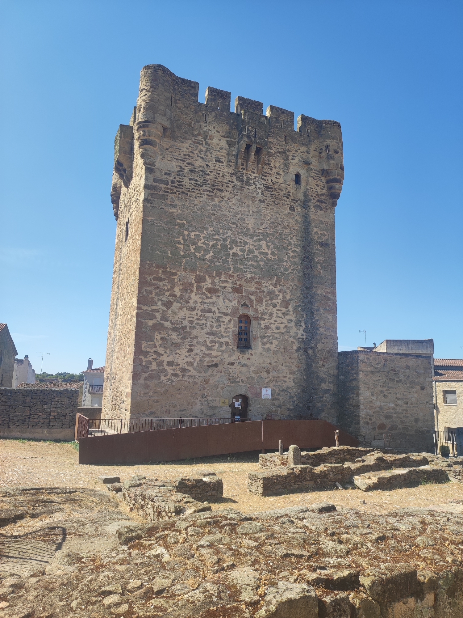 The image shows a close-up view of the Torre de la Calzada, a historic stone tower located in the city of Sobradillo, Spain. The tower is a tall, imposing structure with a crenellated top and small windows. It is built of rough-hewn stone blocks of varying sizes and colors, giving it a rustic and ancient appearance. The tower stands against a clear blue sky. In the foreground, there are visible ruins of stone structures, suggesting an archaeological site surrounding the tower. A metal ramp leads to the entrance of the tower. Several other buildings, constructed from stone and tile, can be seen in the background, indicating the tower is situated within a town or village. The ground is covered in dry, light-colored soil and scattered stones. There are no people visible in the image.