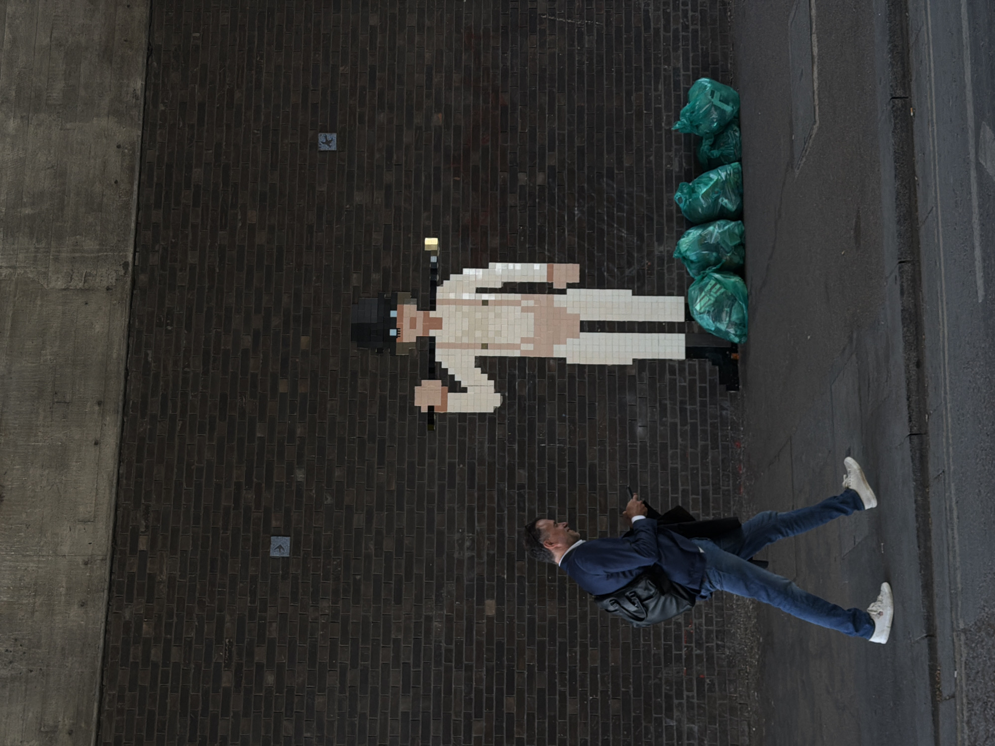 The scene, viewed from above, is a London street corner. A man in a blue suit, jeans, and white sneakers is walking along the sidewalk, looking at his phone. To his right, against a brick wall, is a pixelated mosaic resembling Alex DeLarge from "A Clockwork Orange." Next to the mosaic are four green trash bags. A gray concrete wall is visible to the left. Two small square markers are embedded in the brick.