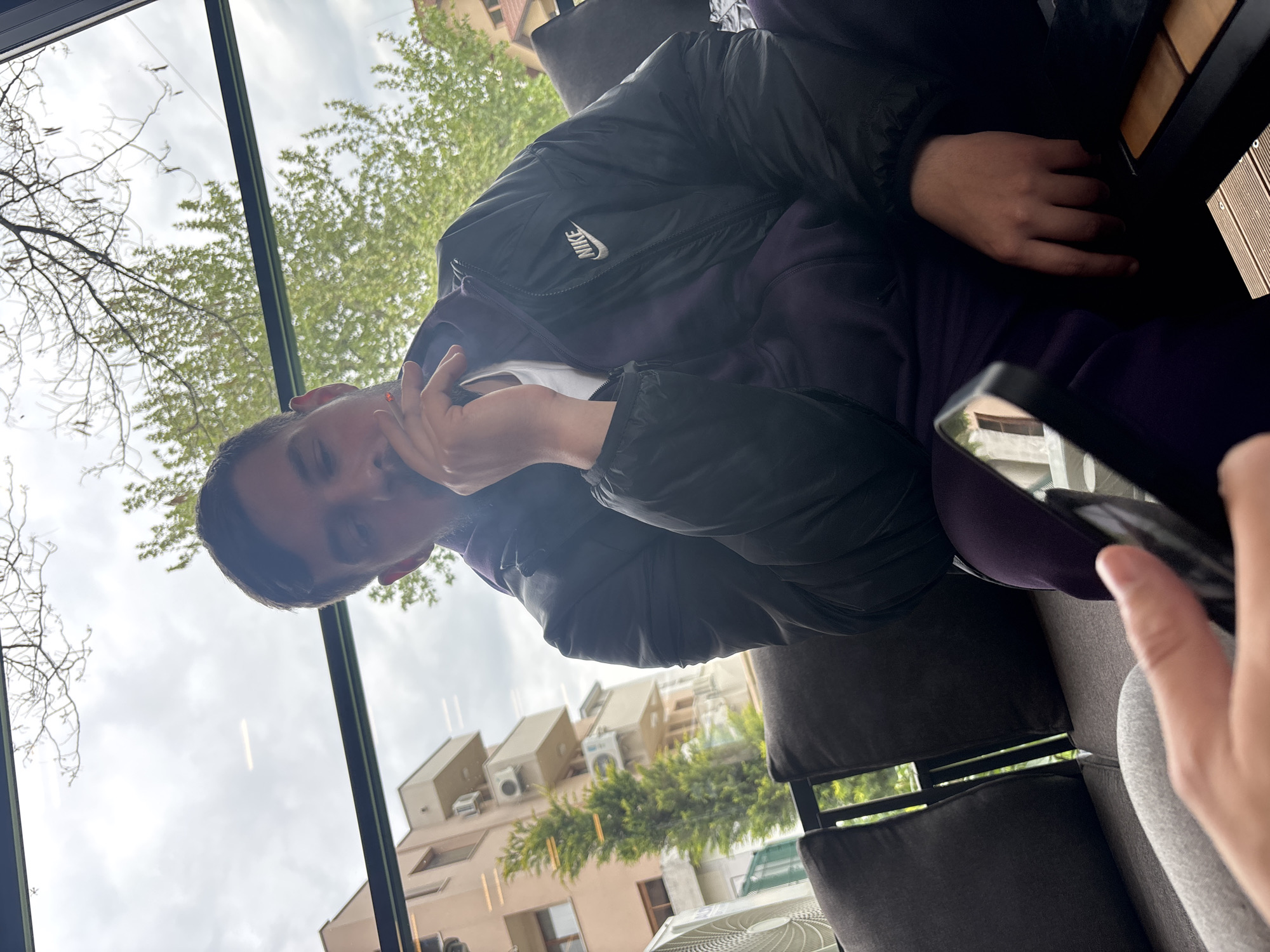 A person wearing a black Nike jacket and purple pants sits in a dark gray chair near a window. The view from the window includes buildings, trees, and a cloudy sky. The image's location is inferred to be Plovdiv, Bulgaria, but this is not directly evident in the image itself. The person holds a compact makeup case in their left hand. A table is next to the person.