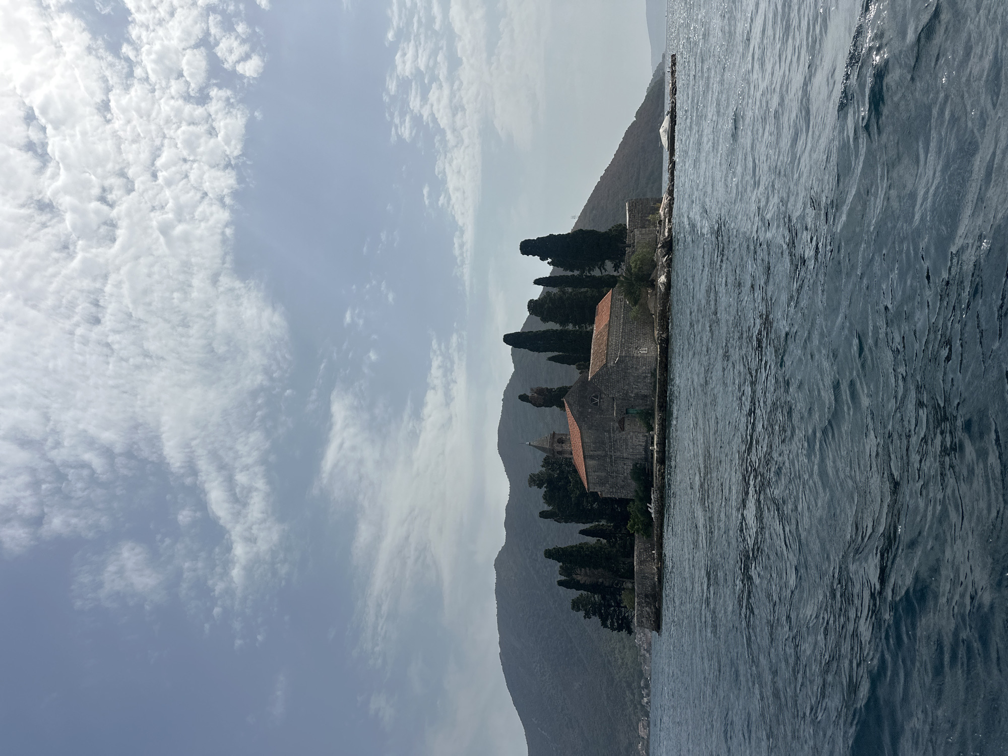The image captures the islet of Our Lady of the Rocks (Gospa od Škrpjela) in the Bay of Kotor, near the town of Perast, Montenegro. The islet, a man-made island, is dominated by a stone church with a red tiled roof and a bell tower. Several tall, slender cypress trees are planted around the church, and a low stone wall encloses the islet. The sea in the foreground is dark blue and moderately choppy, with visible ripples and a slight wake on the right side of the frame. In the background, a steep, green-covered mountain slopes down to the water, with a hint of buildings visible on the distant shoreline. The sky is partly cloudy, with patches of blue sky interspersed with white, wispy clouds. The overall impression is one of a serene, historic island situated in a beautiful natural setting.