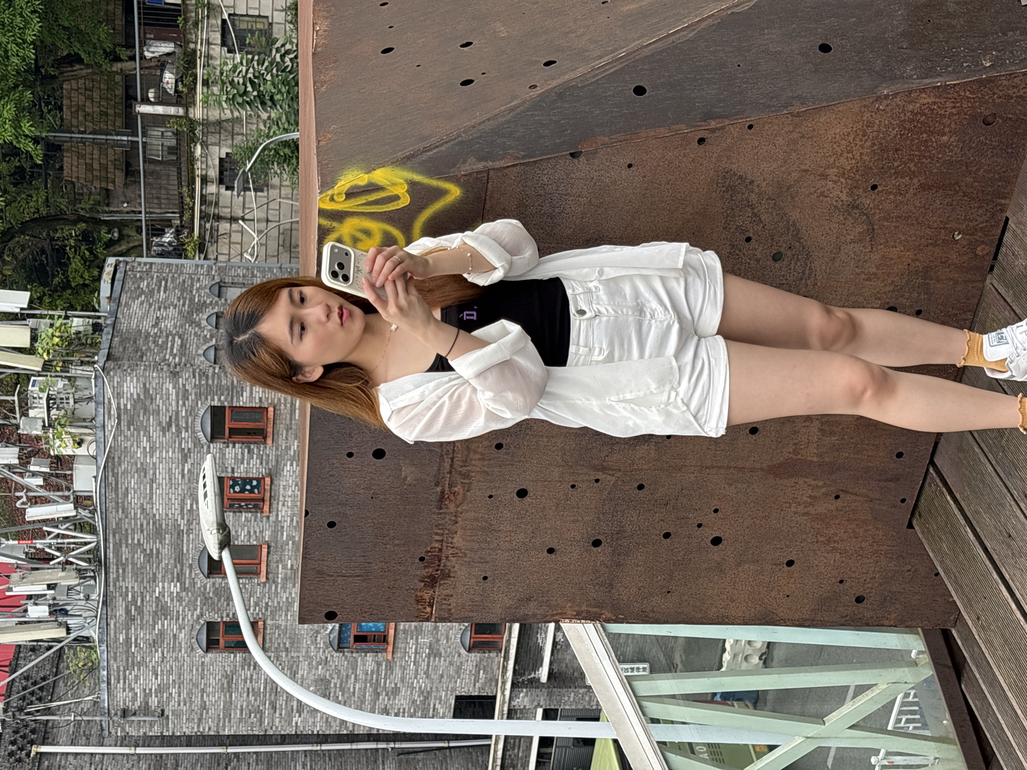 A young woman is lying on a weathered steel structure in Yuzhong District, China, operating a smartphone. She wears a black top, white shorts, and a white sheer button-up shirt, along with white sneakers and a gold-colored anklet. The steel structure, marked with holes and yellow graffiti, intersects with wooden planks. The background reveals a cityscape with brick buildings, street lights, and what appears to be a green bridge structure.