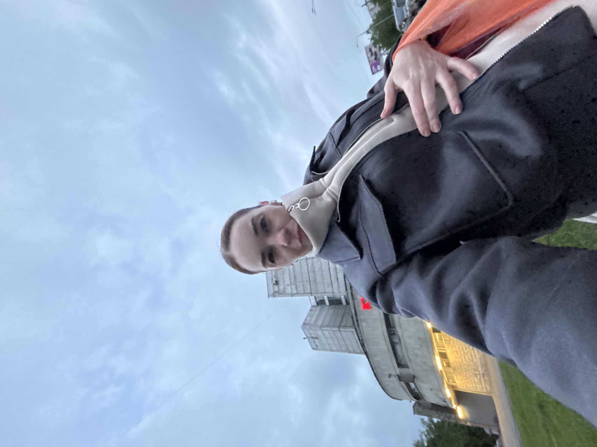The image depicts a person with light skin and short brown hair, wearing a black jacket over a beige hooded sweatshirt. The person is looking towards the camera and smiling. Behind them, a building with a curved facade and a tower-like structure is visible. There are lights illuminating the building. The sky is cloudy and gray. The location is in the city of Unknown, Unknown.