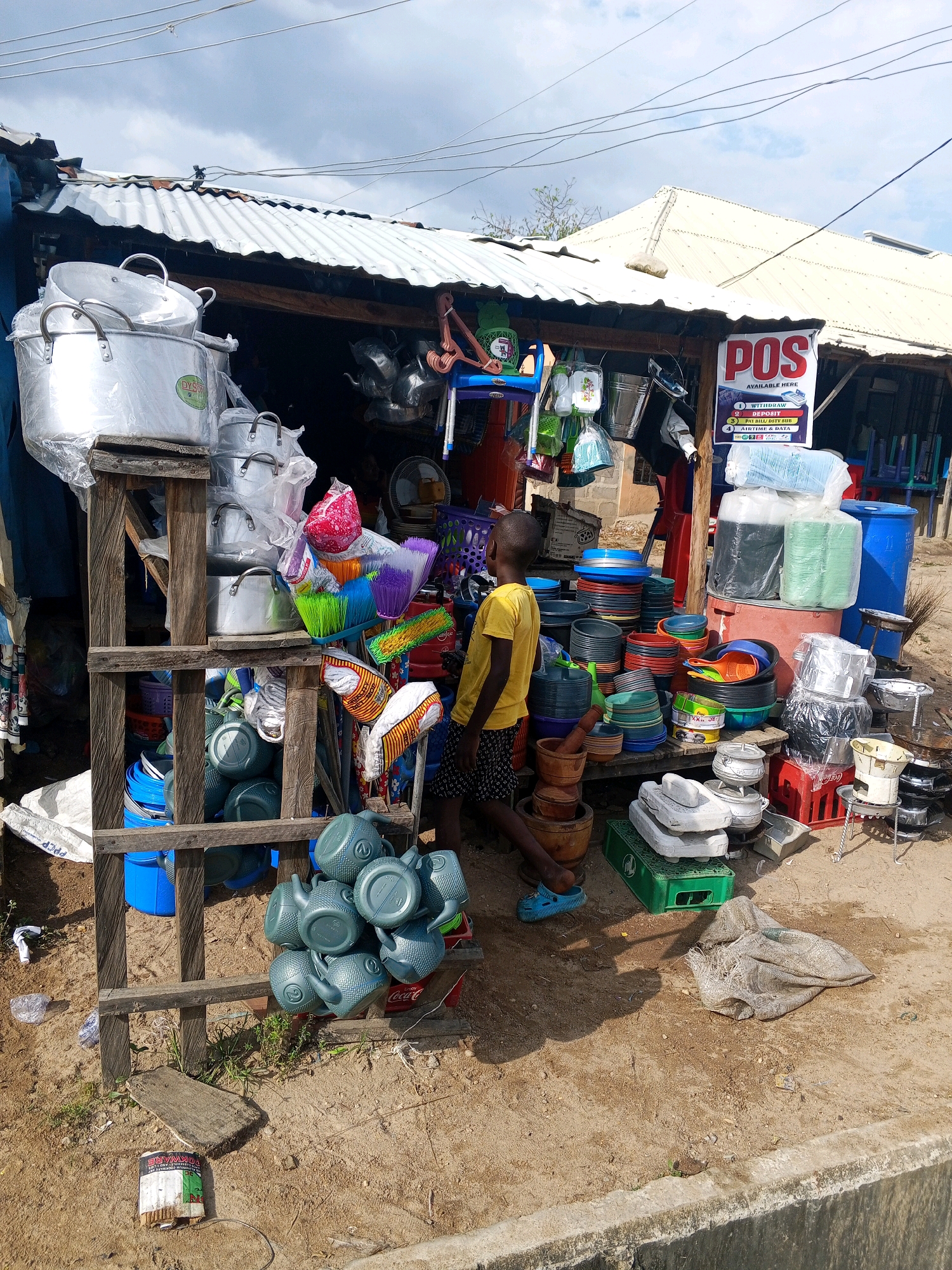 In Zariagi, Nigeria, a bustling open-air market stall displays a variety of kitchenware and household items. A young boy, wearing a yellow t-shirt and black shorts, stands in front of the stall, looking towards the left. The stall, constructed with corrugated metal roofing and wooden supports, is overflowing with merchandise. Prominently displayed are large aluminum pots, stacked on wooden crates. To the right, a "POS AVAILABLE HERE" sign advertises financial services. Various colored plastic basins, buckets, and utensils are neatly arranged. In the foreground, several grey watering cans are piled up. The ground is sandy and uneven, with some scattered debris and vegetation. The scene is bathed in natural daylight, with shadows indicating an overcast sky.