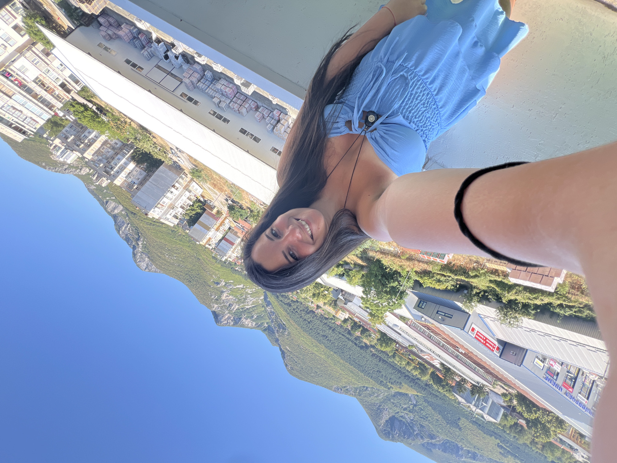 A woman with long, dark hair takes a selfie from a balcony overlooking a cityscape with mountains in the background. She is wearing a blue strapless dress with ruching on the bodice and a black necklace with a circular pendant. Her right arm is extended, holding the camera. The background shows a large industrial building with stacks of materials outside, residential buildings, trees, and a lush green mountain range under a clear blue sky. The location is Vratsa, Bulgaria.