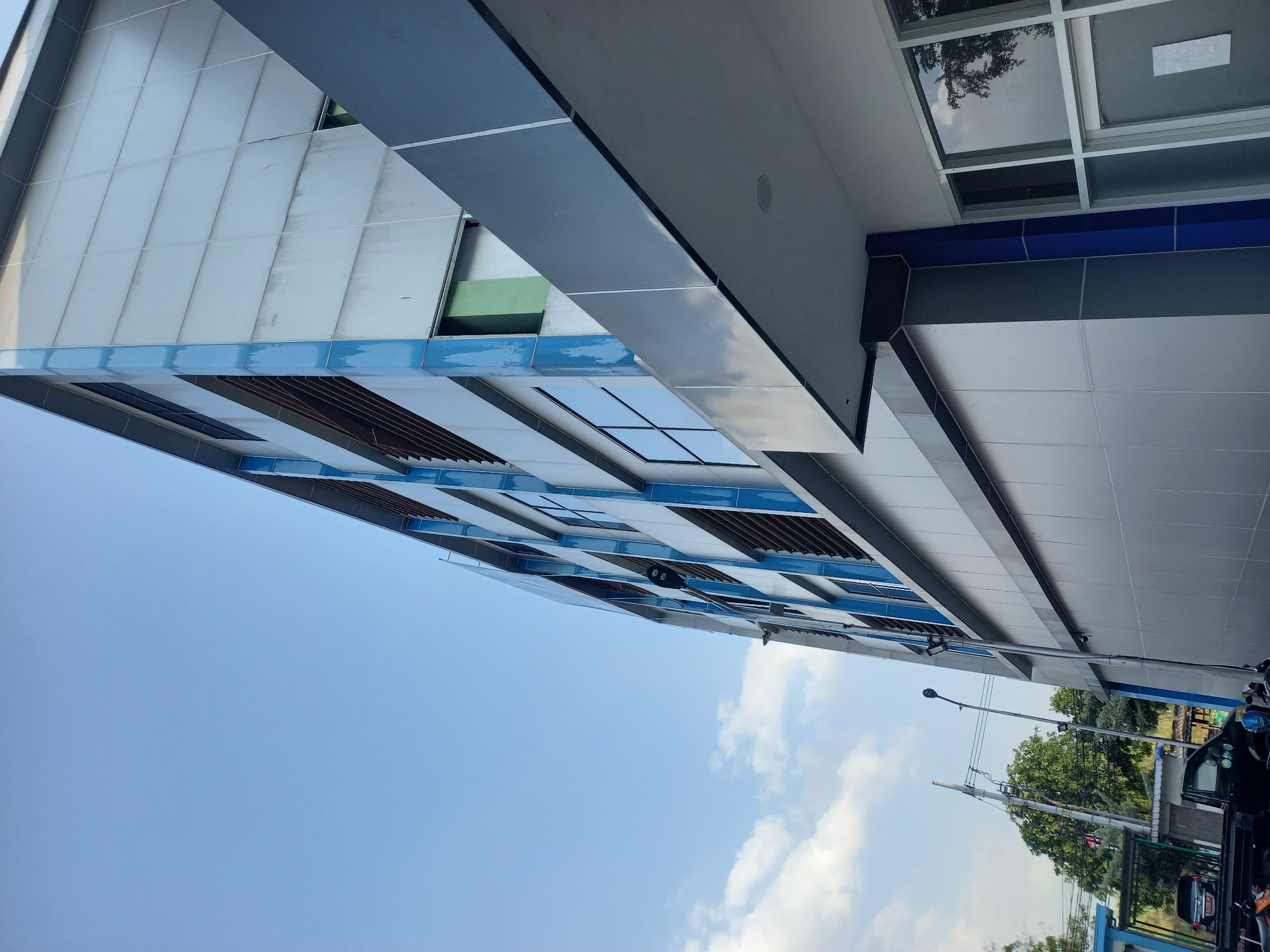 The image shows the exterior of a modern, multi-story building with a light-colored metallic facade and blue accents. The building has numerous windows and slatted vents. In the foreground, part of an overhang with recessed lighting is visible. The sky above is clear blue with a few scattered clouds. To the left of the building, there are trees, utility poles, streetlights, and a few vehicles including a black pickup truck and a motorcycle. The scene is likely in an urban or semi-urban area.