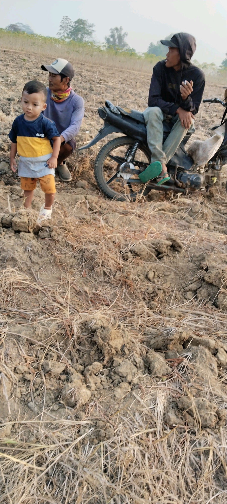 In Tanjung Ratu Ilir, Indonesia, a young boy stands in a dry, tilled field. He wears a shirt with yellow and blue stripes and orange shorts. To his left, an adult man wearing a blue long-sleeved shirt and a cap squats, looking down at the boy. Behind them, another individual is seated on a black motorcycle, wearing a dark hoodie and light-colored pants. This person is holding a cigarette and looking to the right. The ground is covered in dry soil and scattered straw. In the background, a hazy expanse suggests a treeline under a bright sky.