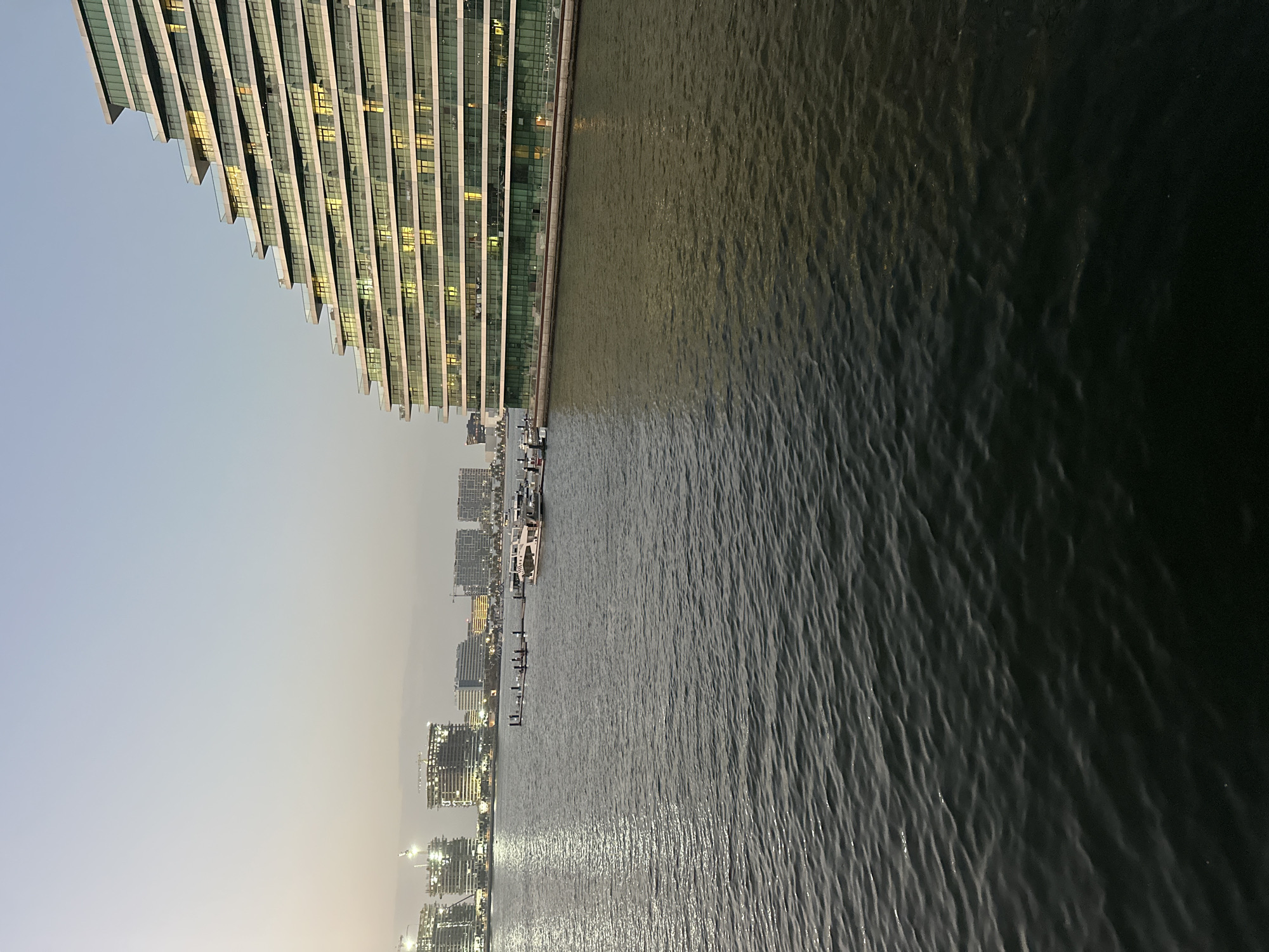 The image shows a view from the water in Abu Dhabi, United Arab Emirates. The left side of the image depicts modern buildings and skyscrapers, with a prominent structure featuring a unique saw-tooth edge design along its roofline. Lights emanate from within the building, suggesting it is evening or night. The right side of the image is dominated by the dark and rippled water. In the middle ground, there are glimpses of more buildings lining the shore, along with boats docked at the pier. The sky is a pale, dusky color, indicating either sunrise or sunset.