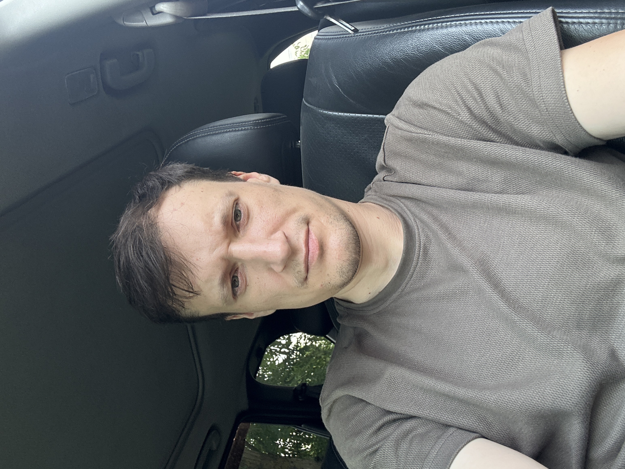 The image is a selfie taken from inside a car. A man with short, dark hair and a light complexion is sitting in the driver's seat. He is wearing a grey t-shirt. His expression appears concerned or serious. The interior of the car is visible, including a black leather seat and the sunroof. Through the windows, trees and greenery are visible, suggesting an outdoor setting, possibly near a rural or suburban area. The image is lit by natural daylight. The man is positioned in the center of the frame, with the car's interior filling the rest of the image. There is no indication of the location being the city of Makiv, Ukraine.