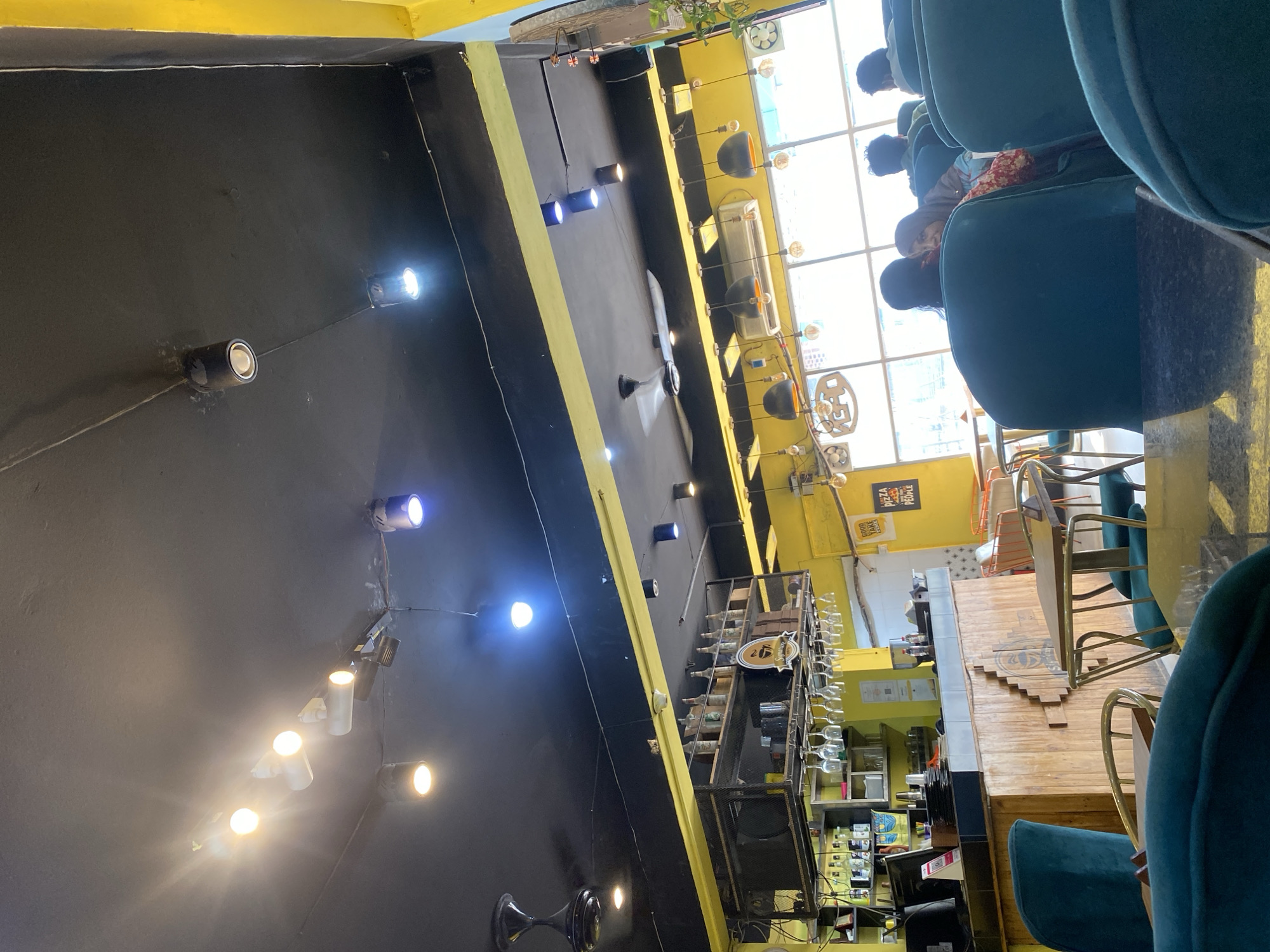The image depicts the interior of a cafe with a yellow and black color scheme. The ceiling is black with multiple track lights and pendant lights. A black ceiling fan is visible on the left. The walls are predominantly yellow, with a section of wooden paneling behind a bar area. The bar area has shelves stocked with bottles and glasses hanging above it. In the foreground, several teal-colored chairs are arranged around tables. In the background, through large windows, daylight is visible, and several patrons are seated, some facing away from the camera and others partially visible. One woman is looking towards the camera. The overall ambiance suggests a contemporary cafe or restaurant setting, possibly in Dhaka, Bangladesh, given the context of previous image analyses.