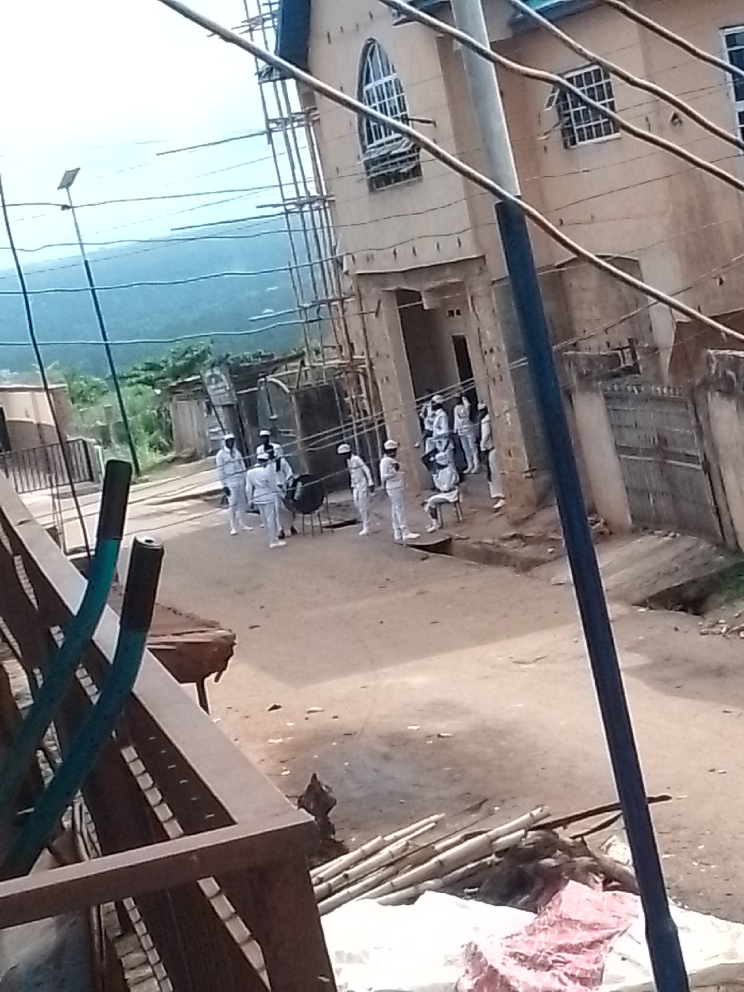 A street scene in Unknown, Unknown, shows a group of approximately ten individuals in white uniforms and hats gathered near a building under construction. Scaffolding is visible on the building's facade. The foreground includes what appears to be construction debris and metal framing. In the background, there are mountains and vegetation. Overhead power lines crisscross the scene. The overall lighting suggests it is daytime.