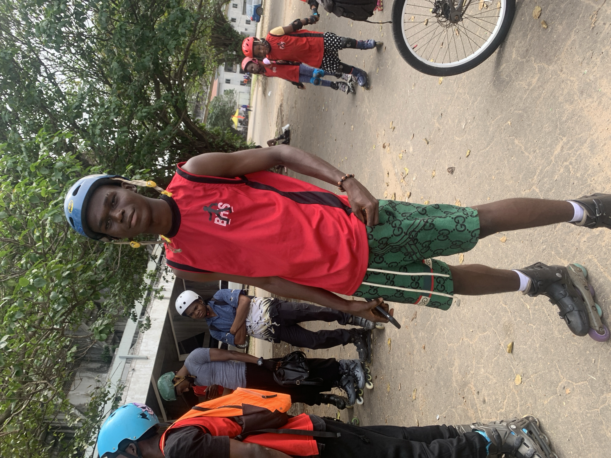 In Alagbede, Nigeria, a group of individuals are gathered outdoors, seemingly preparing for or engaged in rollerblading activities. The focal point is a young male standing in the foreground, wearing a red sleeveless shirt, green patterned shorts, and black rollerblades. He is also wearing a blue helmet with yellow embellishments. To his left, another male in a red vest and a blue helmet is partially visible. Behind them, several other individuals are present, including a man in a grey t-shirt and black pants, and another wearing a blue shirt and a white helmet. In the background, two young girls in red shirts and helmets are also on rollerblades, along with other figures and a vehicle. The environment appears to be an urban or semi-urban outdoor setting, with paved ground, trees, and buildings visible in the distance. The overall atmosphere suggests a recreational gathering focused on rollerblading.