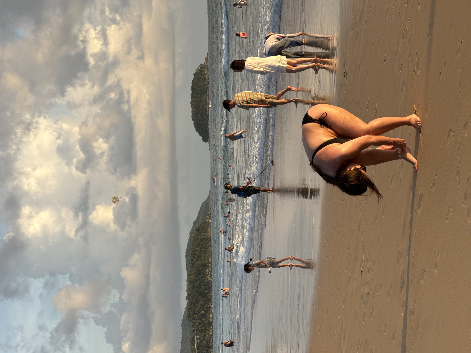 This is a photograph of Karon Beach in Karon, Thailand, at sunset. The sky is filled with dramatic clouds, tinged with the warm colors of the setting sun. In the distance, a parasail is visible against the cloudy sky. The ocean has gentle waves breaking on the shore, and several people are visible in the water, swimming and wading. A small, tree-covered island is visible in the middle distance.
In the foreground, a woman in a black bikini is bending over, appearing to pick something up from the sand. Her back is to the camera. To her right, a man in a striped shirt and shorts walks towards the water, looking out at the ocean. To his right, a woman in a white outfit stands with her back to the camera, looking at the ocean. Further to the right, a person wearing a grey shirt and dark pants is bent over, seemingly searching the sand. On the wet sand, reflections of people and the sky are visible.
Further back on the beach, a young girl in a patterned dress stands near the water's edge, holding a small object. Several other people are scattered along the shoreline and in the water. The sand is golden brown, with footprints visible in places. The overall scene is one of a relaxed, late afternoon at a popular tourist beach.