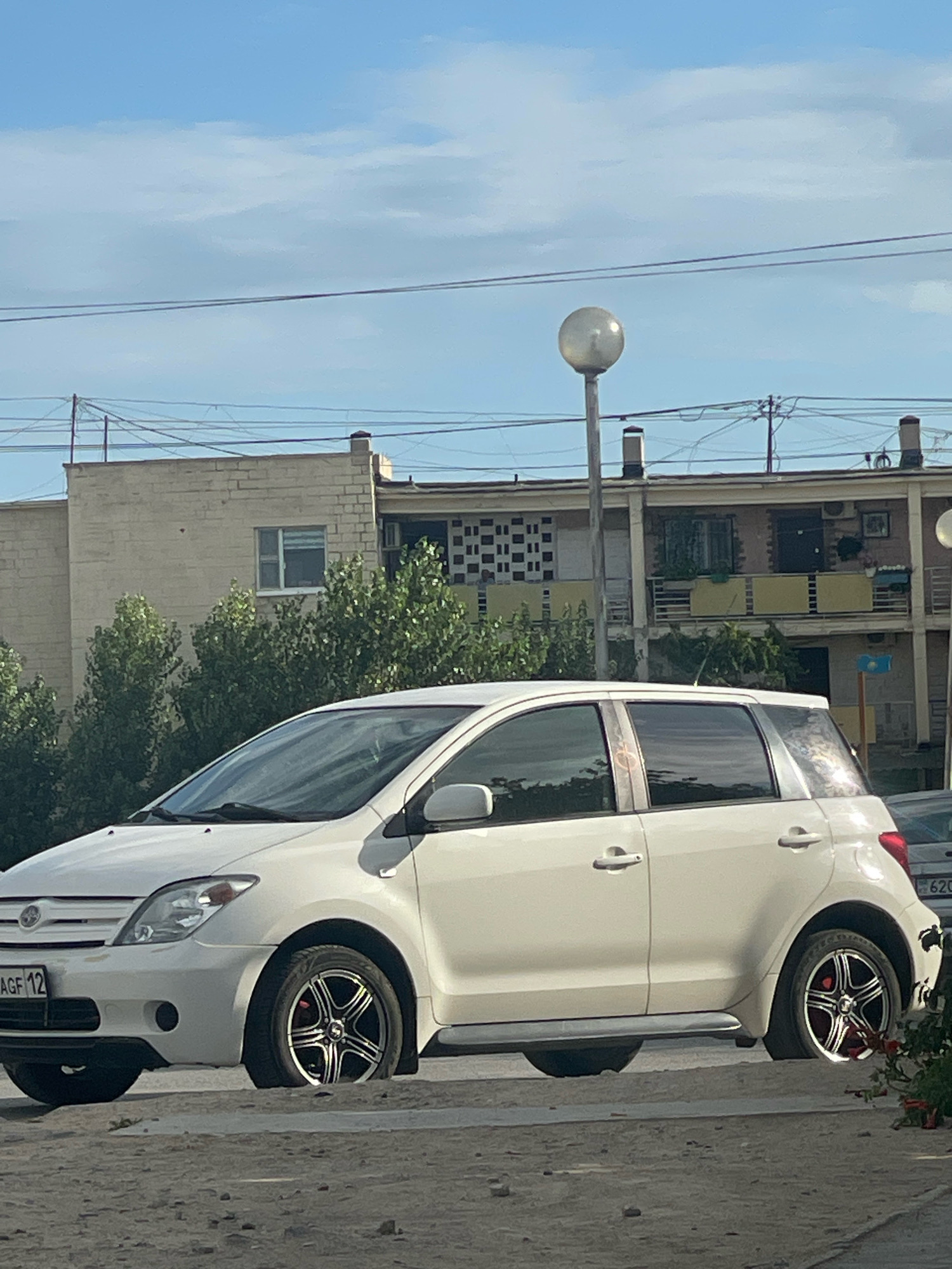 A white Toyota Ist hatchback is parked on a dirt road. The car has custom rims and a flat tire on the front passenger side. In the background, there is a multi-story building with balconies and windows, as well as trees and overhead power lines. The sky is blue with scattered clouds. The license plate is partially visible and appears to read "AGF 12". Another license plate is partially visible on the rear of the car, with the numbers "620". The scene appears to be in the city of Aktau, Kazakhstan.