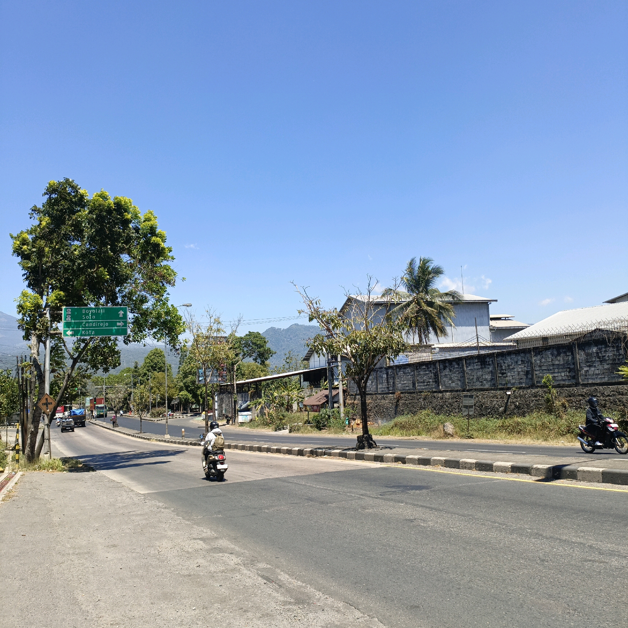 The image depicts a road in Salatiga, Indonesia, on a clear, sunny day. A large green signpost stands on the left side of the road, indicating directions to Boyolali, Solo, Candiirejo, and Kota. A large tree with lush green foliage is situated next to the signpost. Several vehicles, including cars, trucks, and motorcycles, are present on the road, some moving and others parked. Buildings and a stone wall line the right side of the road, with some greenery and trees interspersed. In the background, a mountain range is visible. One person is riding a motorcycle with a beige backpack, heading away from the camera. Another person is on a motorcycle closer to the camera, also moving away. The road is paved and appears to be a major thoroughfare. The sky is a vibrant blue with no clouds.