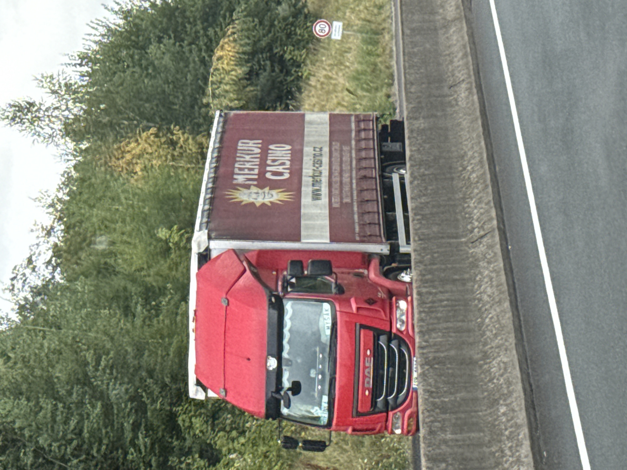 The image captures a red DAF truck with a white trailer driving on a highway. The trailer is branded with "MERKUR CASINO" and the website "www.merkur-casino.cz". The truck has license plates that appear to read "1SP 4R95". A speed limit sign showing "80" is visible on the right side of the road. The surrounding environment consists of dense green trees and grass, with a concrete barrier separating the highway from the verge. The image does not contain any specific landmarks or features that would place it within the city of Bad Lauterberg im Harz, Germany.