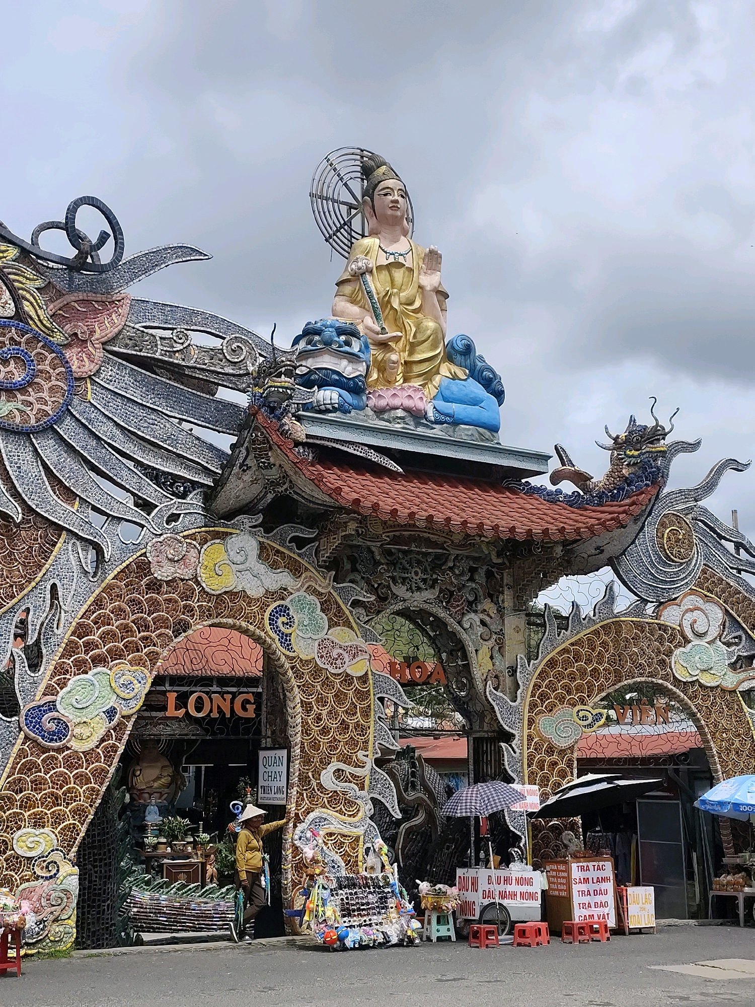 This is a photograph of the entrance to the Long Hoa restaurant in Xuan Thanh, Vietnam. The entrance is elaborately decorated with mosaics of dragons and clouds. Atop the entrance, a large golden statue of Buddha sits on a blue lotus. To the right of the entrance, a woman wearing a conical hat stands near a stall selling drinks. Several umbrellas and small stools are set up in front of the stalls. The sky is overcast.