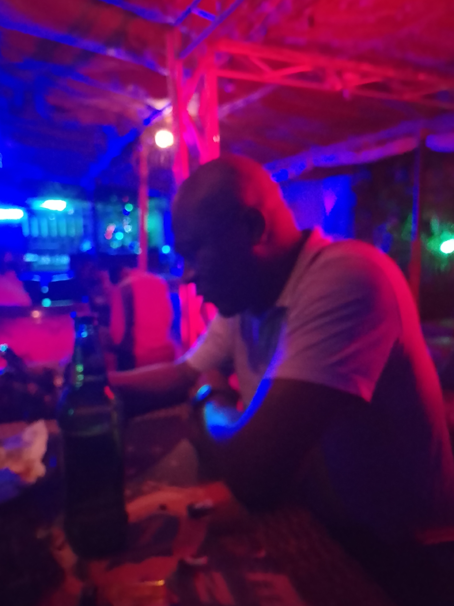 The image shows a person seated at a table in a dimly lit establishment, possibly a bar or lounge. The lighting is primarily red and blue, creating an atmospheric effect. The individual is wearing a light-colored shirt. On the table, there is a bottle of what appears to be beer, alongside other objects. The location is described as being in the city of Unknown, Unknown.
