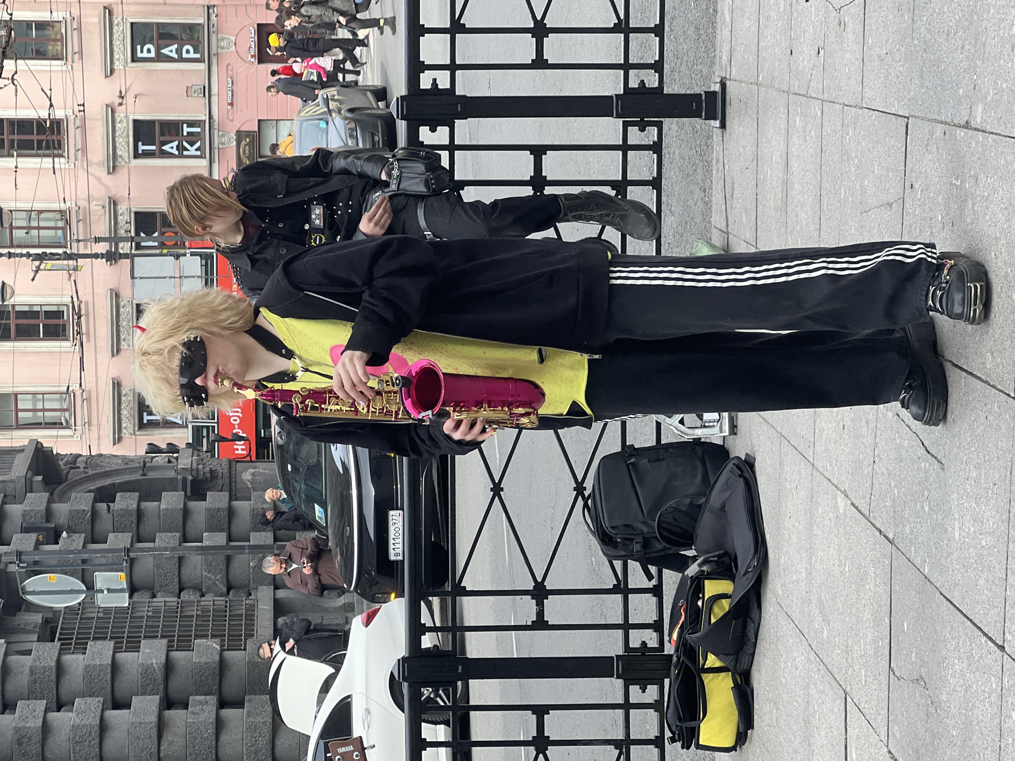 The image captures a street scene in Saint Petersburg, Russia. A performer stands with a pink saxophone near a metal fence alongside a road. The performer is dressed in black clothing with white stripes on the trousers and a yellow top, sporting blonde hair and wearing a spiked eye mask. An individual in a black leather jacket with metal studs sits on the other side of the fence. Cars and pedestrians pass by in the background, and architectural details of buildings are visible. Bags are placed on the ground near the saxophonist. The license plate of a black car reads "B11100 977." Street signs and building names like "HOP-OF" and "БАР" are visible.