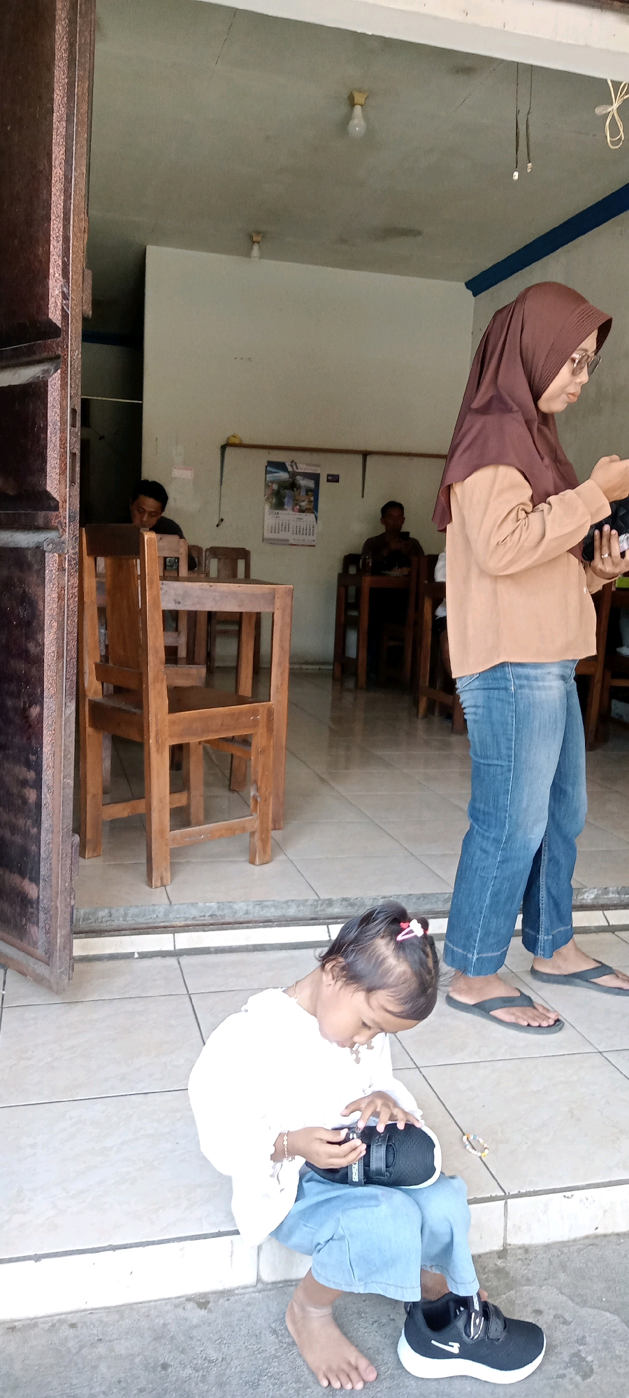 In Desa Karangmojo, Indonesia, a young child with a pink hair accessory is sitting on a tiled floor, holding a black sneaker. The child is wearing a white top and light blue pants. To the right of the child, a woman wearing a brown hijab and glasses is standing, dressed in a tan shirt and blue jeans. She is holding a dark object in her hands. In the background, two men are seated at wooden tables inside a building. One man, with dark hair, is looking down. Another man, further back, is looking at his phone. The environment appears to be an open-air establishment with wooden chairs and tables. Two light bulbs are visible on the ceiling. A calendar hangs on the wall behind the seated men. The entrance to the establishment features a dark wooden frame.