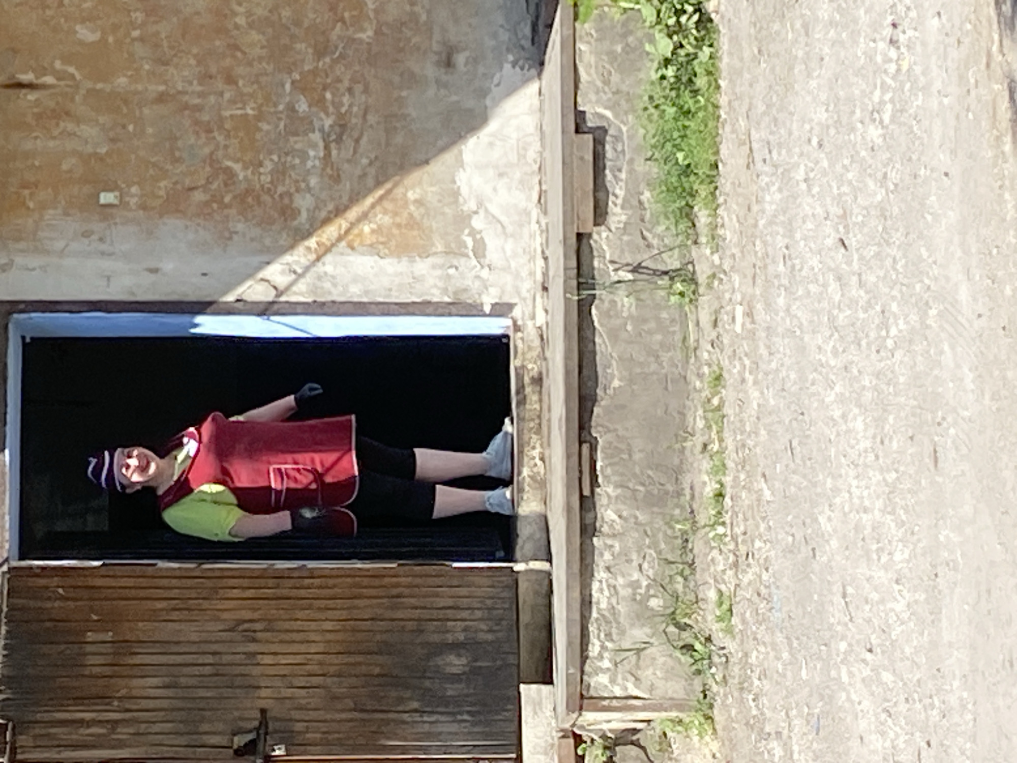 A woman wearing a red apron and a purple and white striped hat stands in the doorway of a building with wooden doors. She has on a light green shirt, black leggings, and light grey shoes. Her hands are gloved. The building has a light brown, textured exterior wall with peeling paint, and a light blue door frame. The doorway is recessed, with a concrete step leading up to it. Vegetation is visible growing on the step and on the ground in front of the building. The ground in the foreground is a gravel or dirt surface. The image appears to be taken in daylight.