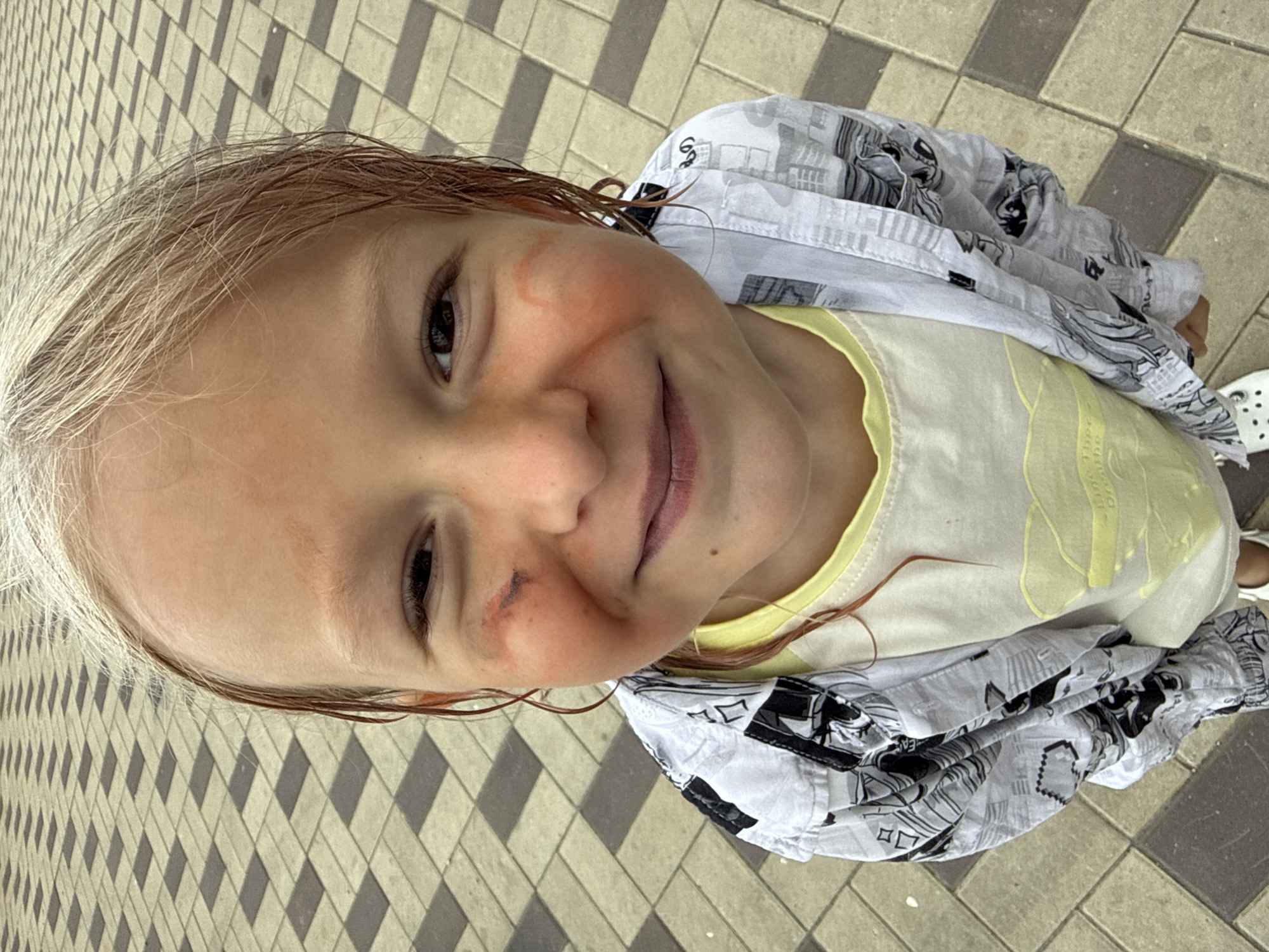 A close-up, high-angle shot shows a young Caucasian child with blonde hair and a slight smile, looking directly into the camera. The child has some reddish marks and a scrape on their left cheek. They are wearing a yellow t-shirt under a grey and white patterned button-up shirt. The background consists of a paved ground with a repeating diamond pattern of light and dark grey tiles.