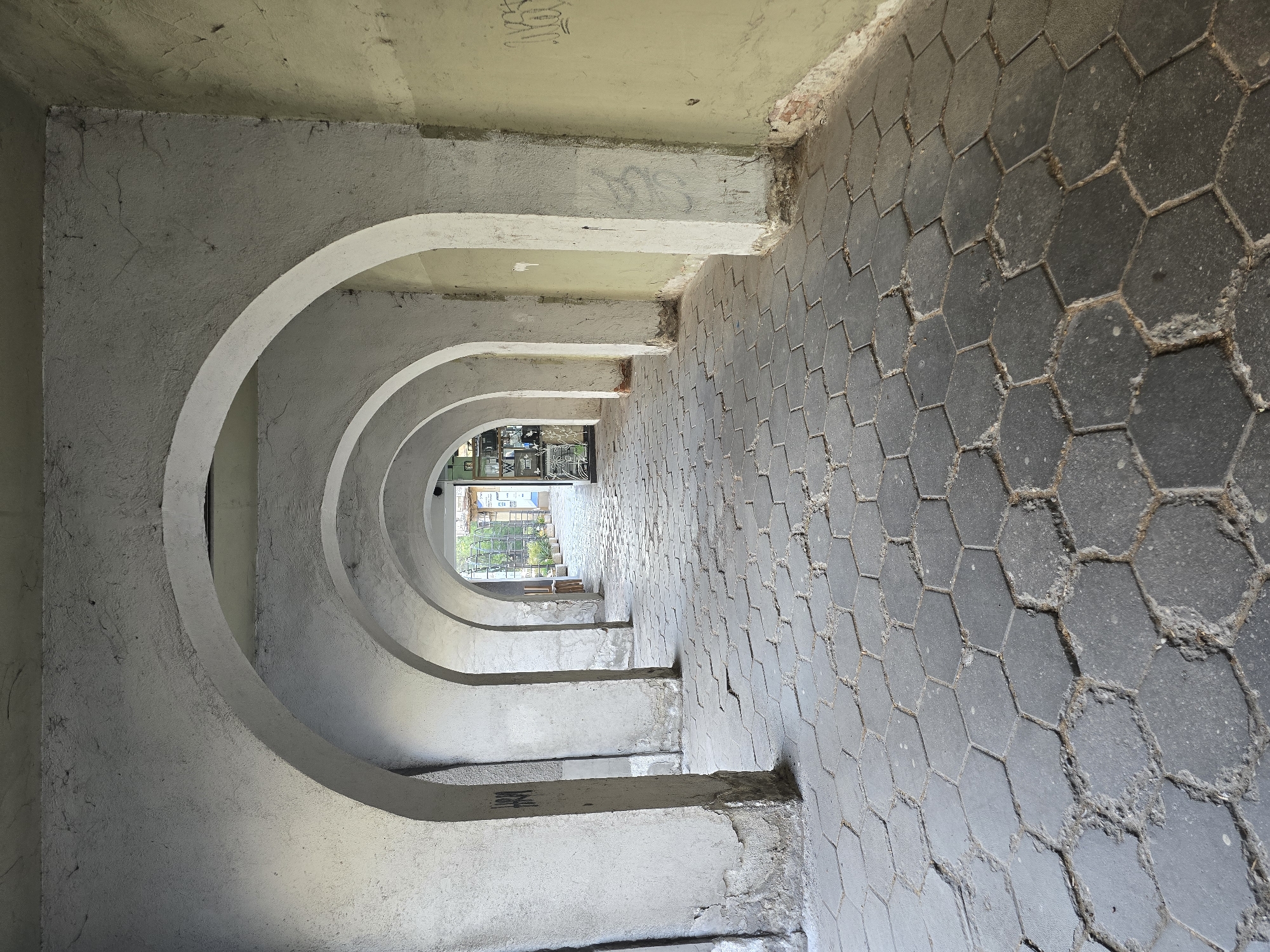 The image depicts a covered passageway in a city location, presumed to be Unknown, Unknown. The passage is defined by a series of repeating, rounded archways on the left side, constructed of concrete. The right side of the passageway consists of a floor paved with hexagonal, dark grey tiles. The passageway appears to be in a state of disrepair, with signs of weathering and deterioration visible on the concrete arches and tiled floor. At the end of the passage, a brighter, outdoor environment is visible through a glazed doorway or opening. The overall perspective is looking down the length of the passageway.
