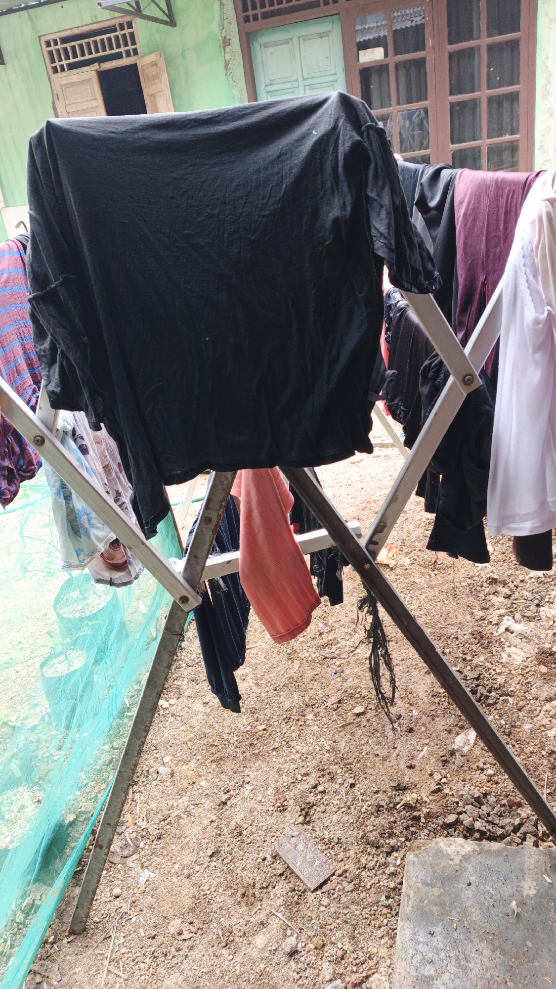 The image depicts a clothes drying rack outdoors in front of a green building with wooden windows. The clothes rack, made of metal, holds various items of clothing, including a large black shirt, a burgundy garment, a white top, and other unidentifiable items. The ground is covered with soil, small stones, and debris, with a green net enclosure visible on the left side. The overall scene suggests a residential area. The specific location is identified as Gunungreja, Indonesia.