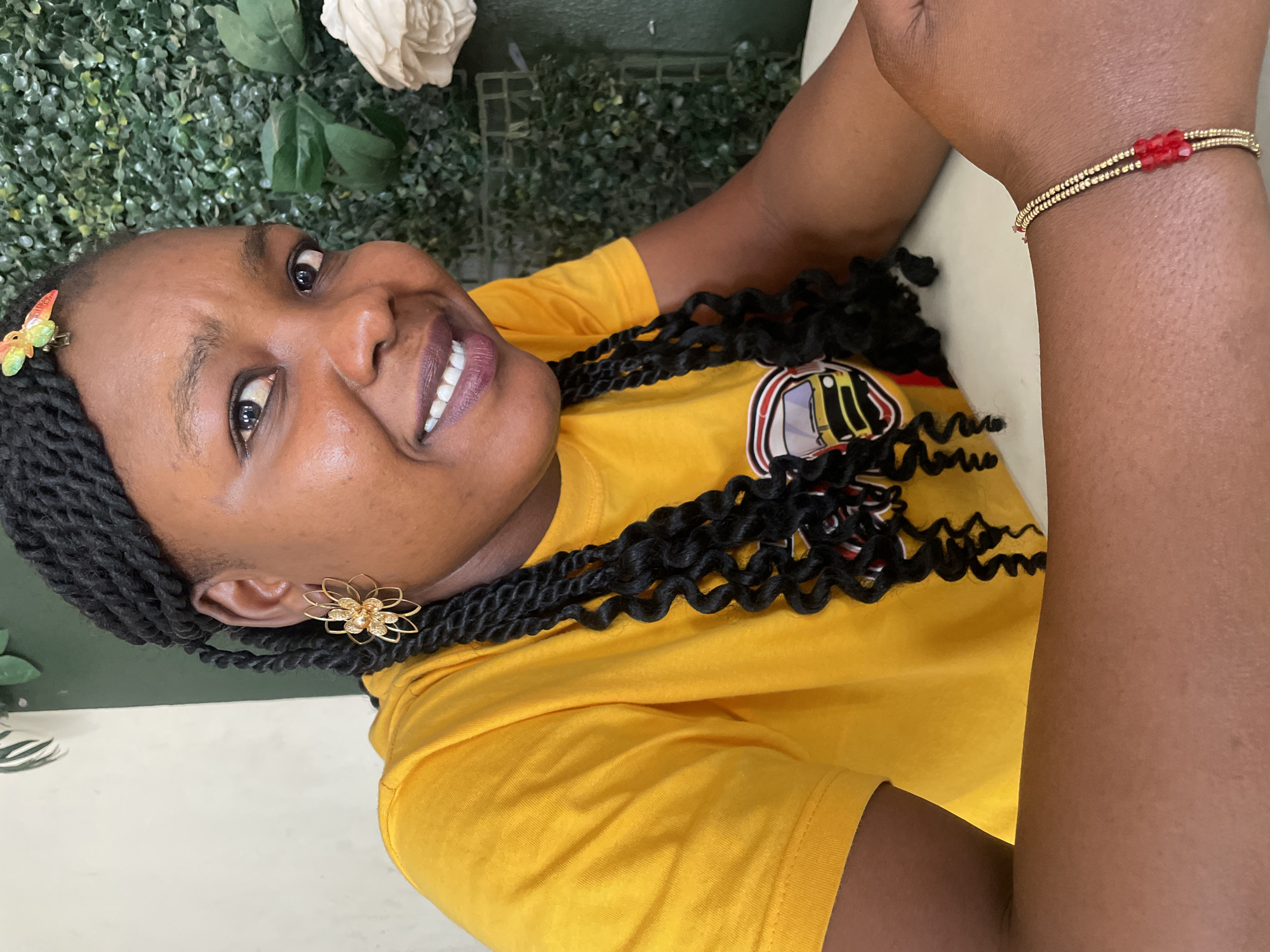 A Black woman is depicted in a cheerful portrait, smiling widely at the camera. Her head is tilted, and she is looking upwards with a warm expression, revealing her teeth. She has dark, braided hair styled with long, wavy extensions, and a small, colorful butterfly clip adorns her hair near her forehead. She is wearing a bright yellow short-sleeved t-shirt with a partially visible graphic logo on the left chest area, featuring white, black, and red elements. A prominent gold-toned floral earring is visible on her left ear. On her right wrist, she wears a delicate bracelet made of gold beads interspersed with small red beads. The woman has dark lipstick on.
The background behind her features a vibrant green wall covered in artificial foliage, resembling a hedge. A white artificial flower is also nestled within the greenery. A light-colored surface, possibly a table or counter, is visible in the foreground where she appears to be leaning. The lighting is soft and even, indicating an indoor setting or a shaded outdoor area during the day in Jos, Nigeria.