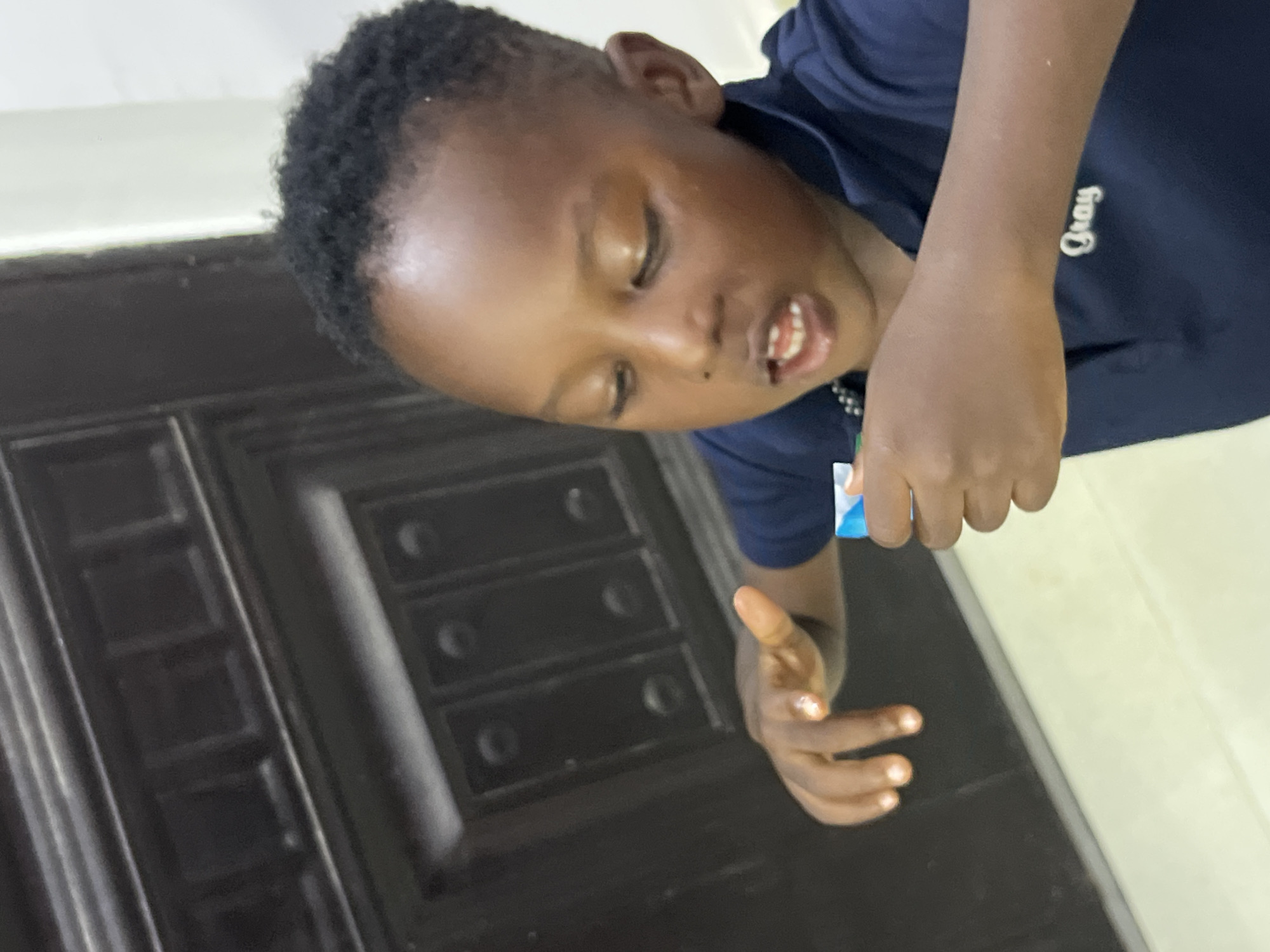 The image shows a close-up, low-angle shot of a young Black boy in Gura Top, Nigeria. He is wearing a dark blue t-shirt with the word "Gray" embroidered in white script on the left side of his chest. The boy is looking downwards with his eyes closed, and his mouth is slightly open, revealing his teeth. His right hand is extended, holding a small, colorful object, possibly a piece of candy or a toy. His left hand is also visible, appearing to reach out or interact with something just out of frame. The background consists of a dark-colored door with raised panels and a lighter-colored wall. The lighting suggests it is daytime, and the overall scene appears to be indoors. The boy's expression could be interpreted as one of concentration or enjoyment.