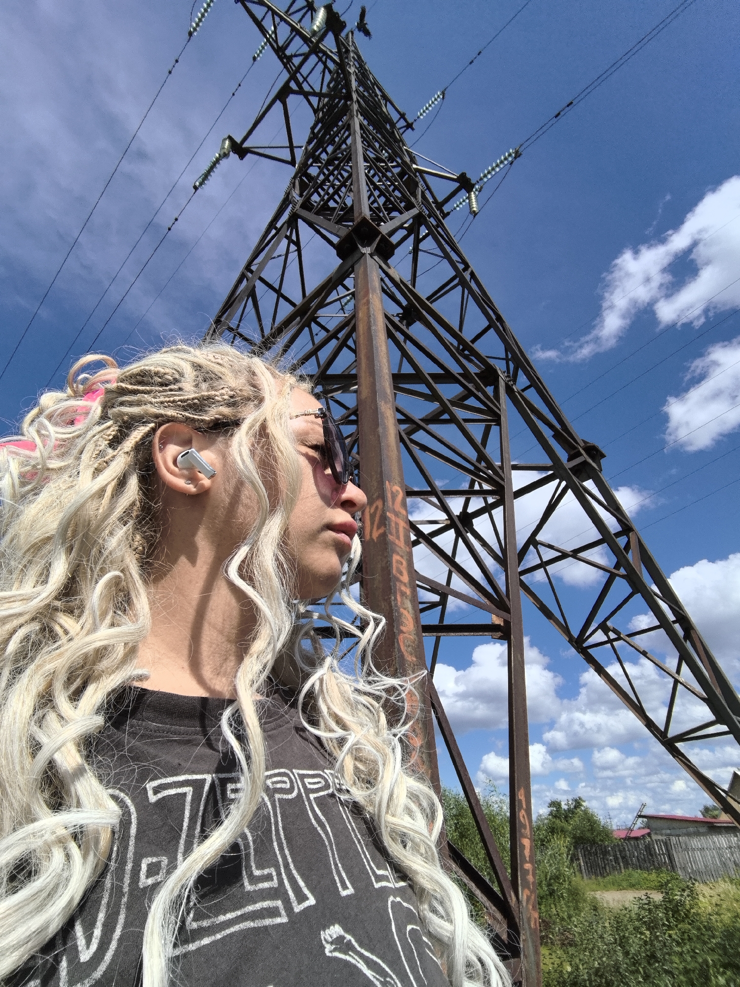 In this media file, a young person with blonde braided hair and pink accents is captured in a low-angle shot, looking up towards a towering electrical pylon against a bright blue sky dotted with fluffy white clouds. The individual is wearing a dark t-shirt with a white graphic, possibly a band logo, and pink-tinted sunglasses. A white wireless earbud is visible in their left ear. The electrical pylon, made of rusted metal, dominates the frame, with thick wires stretching across the sky. The setting appears to be outdoors, with lush green foliage and a wooden fence visible in the background, suggesting a rural or semi-rural location in Noviy, Russia. The lighting indicates it is daytime, likely late morning or early afternoon, with clear and sunny weather. The person's gaze is directed upwards, conveying a sense of contemplation or observation of the imposing structure and the vast sky. The overall mood is one of youthful energy set against an industrial, yet natural, backdrop.