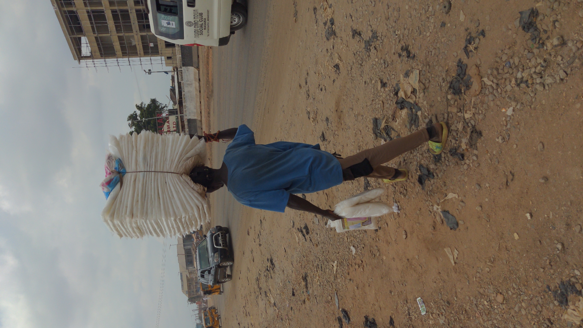 The image shows a man in Ado, Nigeria, carrying a large stack of what appears to be folded fabrics on his head. He is walking on a dirt and gravel area alongside a road. He is wearing a blue shirt and khaki pants, and he is also carrying several plastic bags in his hand. The road has some vehicular traffic, including cars and a truck, and construction equipment can be seen. A building is visible in the background. Litter is scattered on the ground. The text on the side of a white vehicle reads "OGIDI OMO ODUA SOCIAL CLUB."